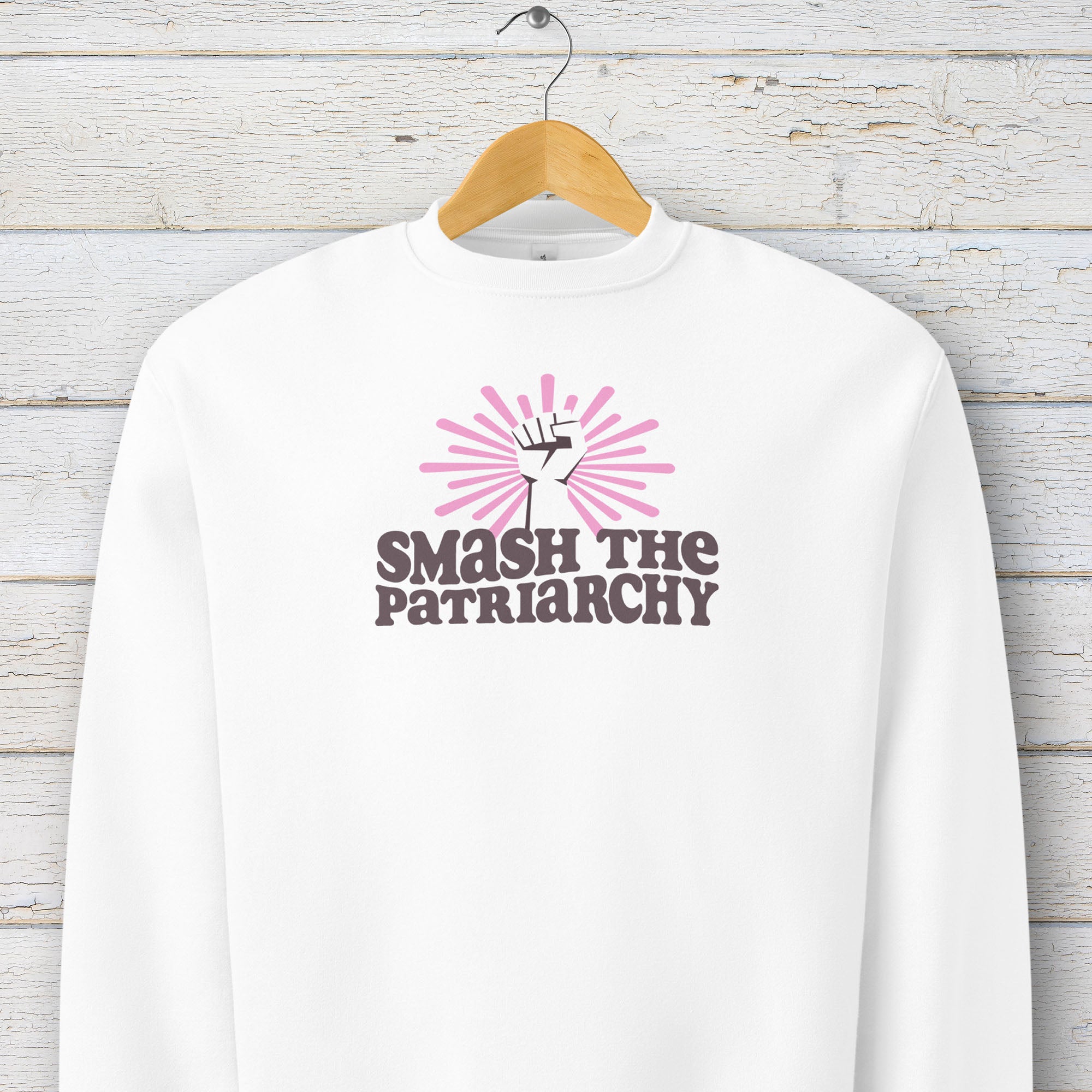 White sweatshirt with a raised fist graphic and pink rays behind it, featuring the text “Smash the Patriarchy.” Hanging on a hanger.