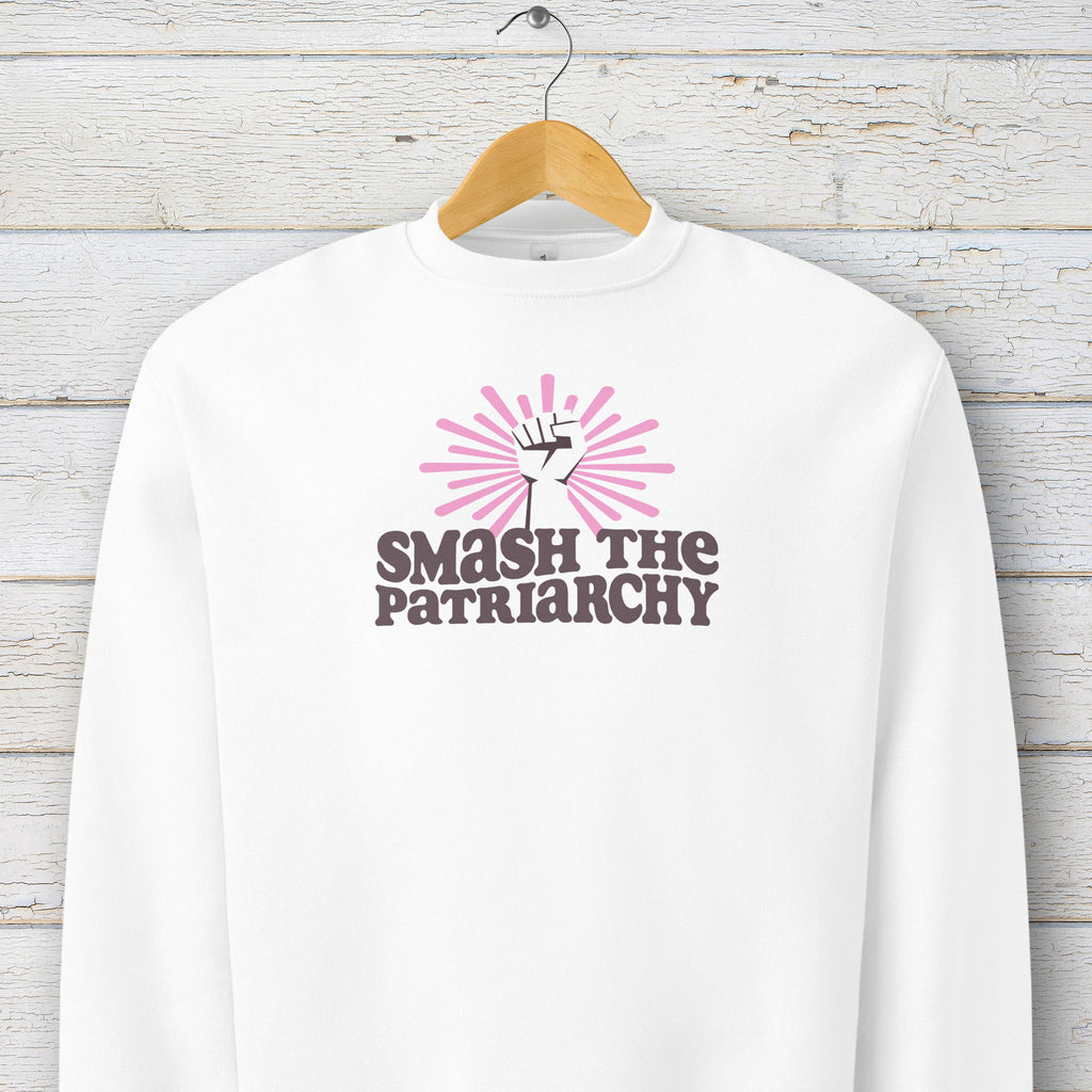 White sweatshirt with a raised fist graphic and pink rays behind it, featuring the text “Smash the Patriarchy.” Hanging on a hanger.