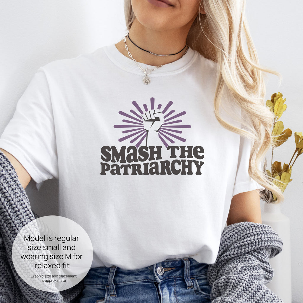Female model wearing white t-shirt with a raised fist graphic and purple burst above the text “Smash the Patriarchy.” Includes size and fit information for the model who is regular size small, and wearing a size M shirt for a relaxed fit.

