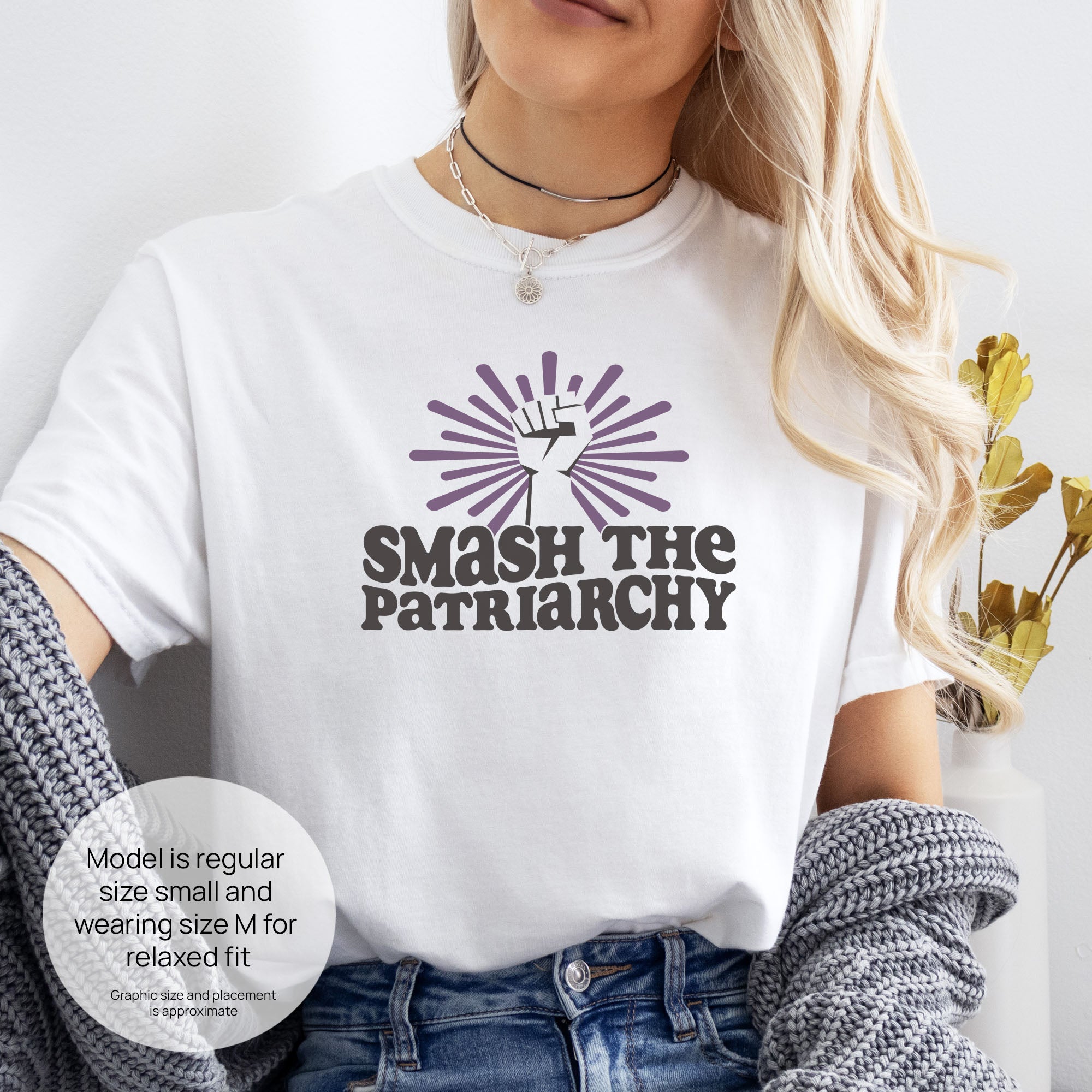 Female model wearing white t-shirt with a raised fist graphic and purple burst above the text “Smash the Patriarchy.” Includes size and fit information for the model who is regular size small, and wearing a size M shirt for a relaxed fit.
