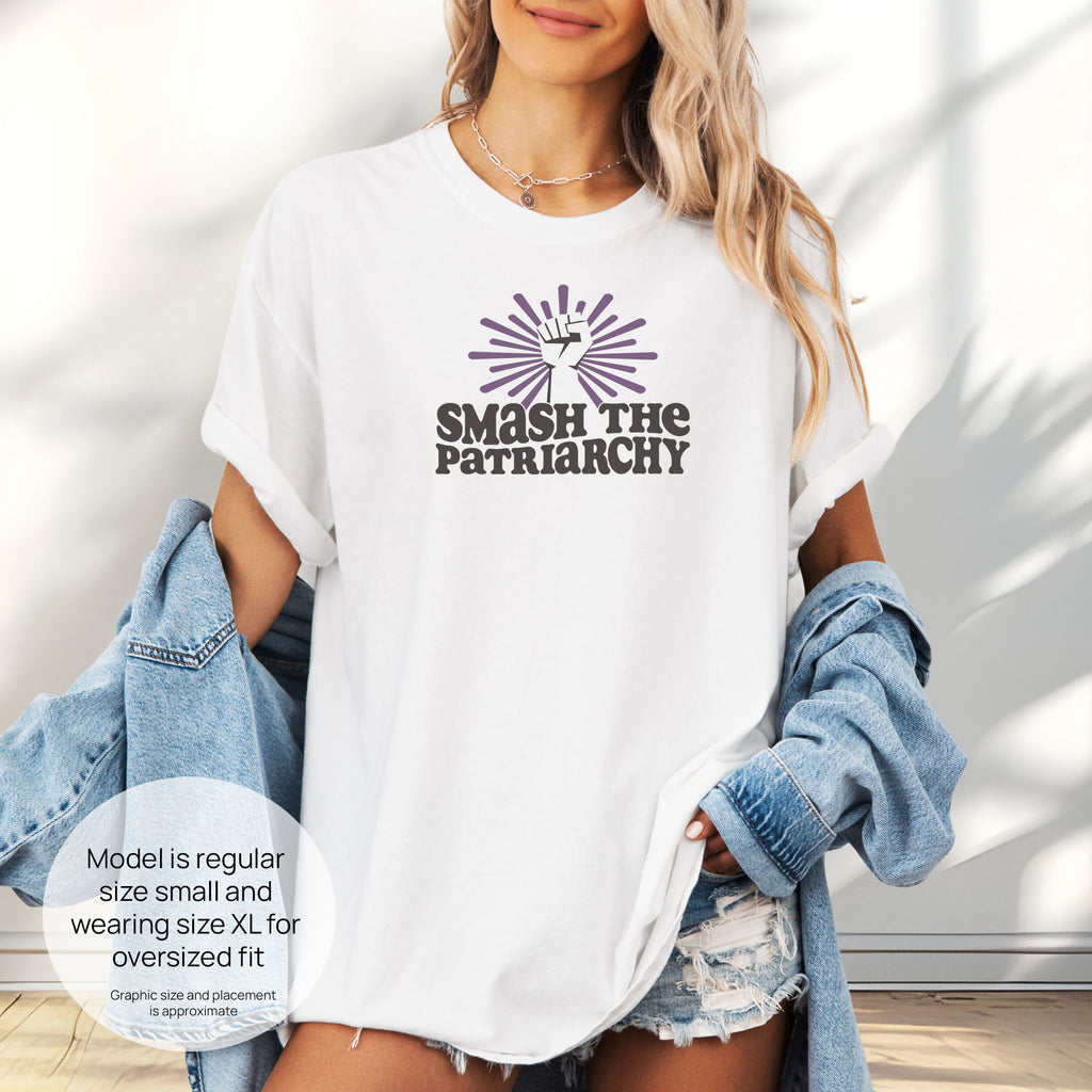 Female model wearing oversized white t-shirt with a raised fist graphic and purple burst above the text “Smash the Patriarchy.” Includes size and fit information for the model who is regular size small, and wearing a size XL shirt for an oversized fit.
