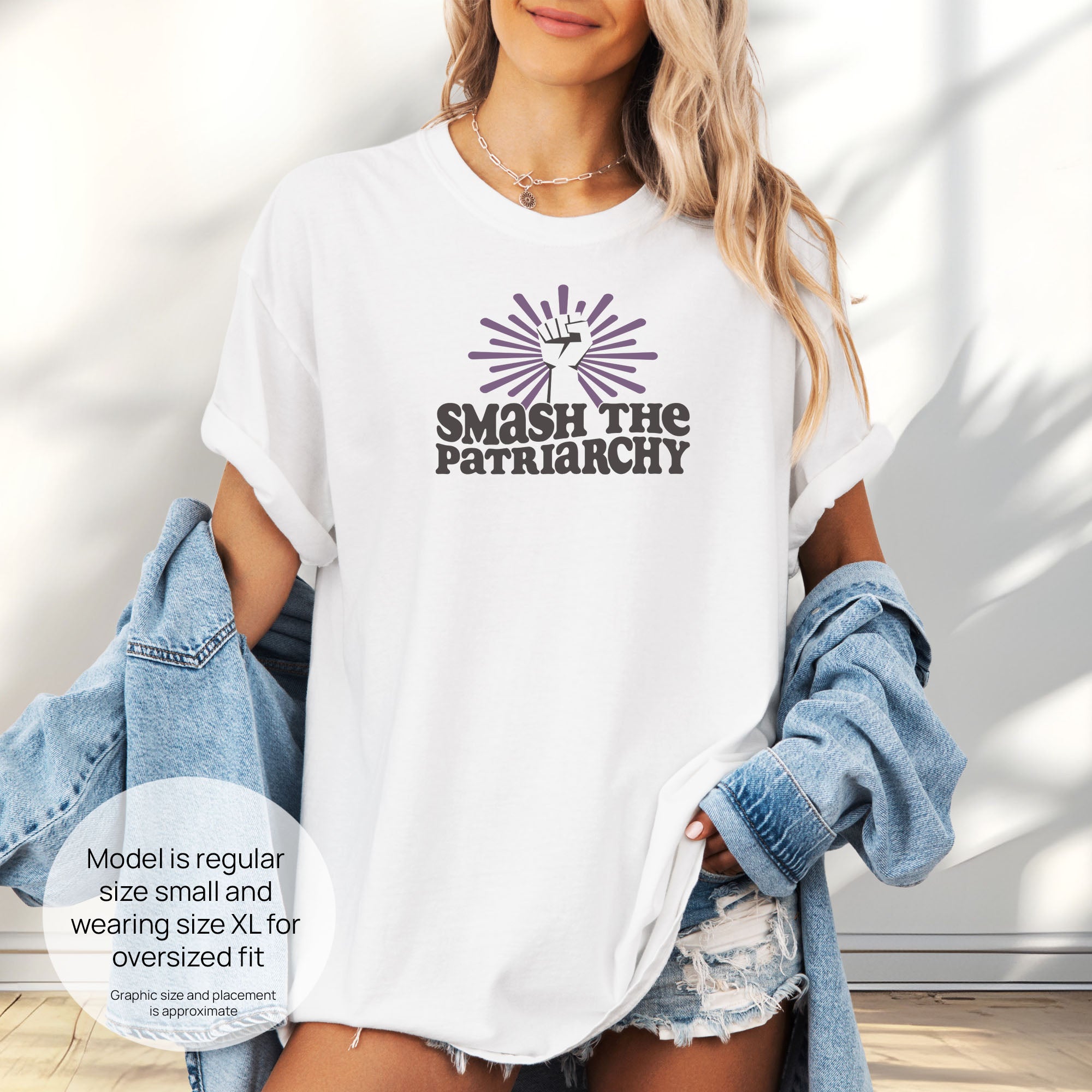 Female model wearing oversized white t-shirt with a raised fist graphic and purple burst above the text “Smash the Patriarchy.” Includes size and fit information for the model who is regular size small, and wearing a size XL shirt for an oversized fit.
