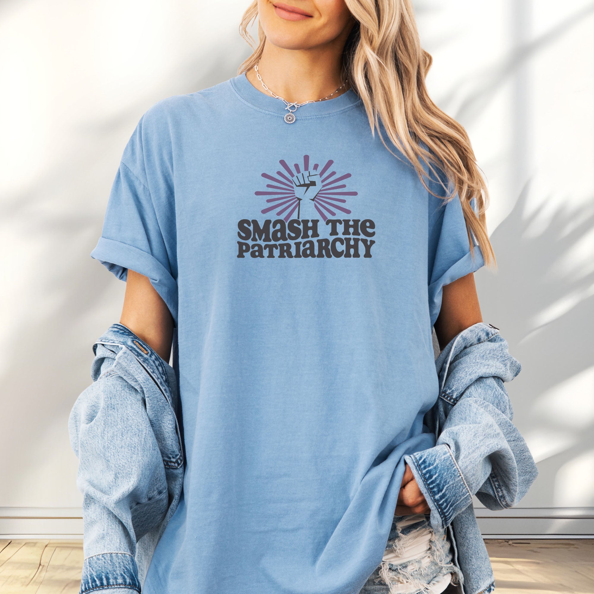Female model wearing oversized washed denim t-shirt with a raised fist graphic and purple burst above the text “Smash the Patriarchy.”