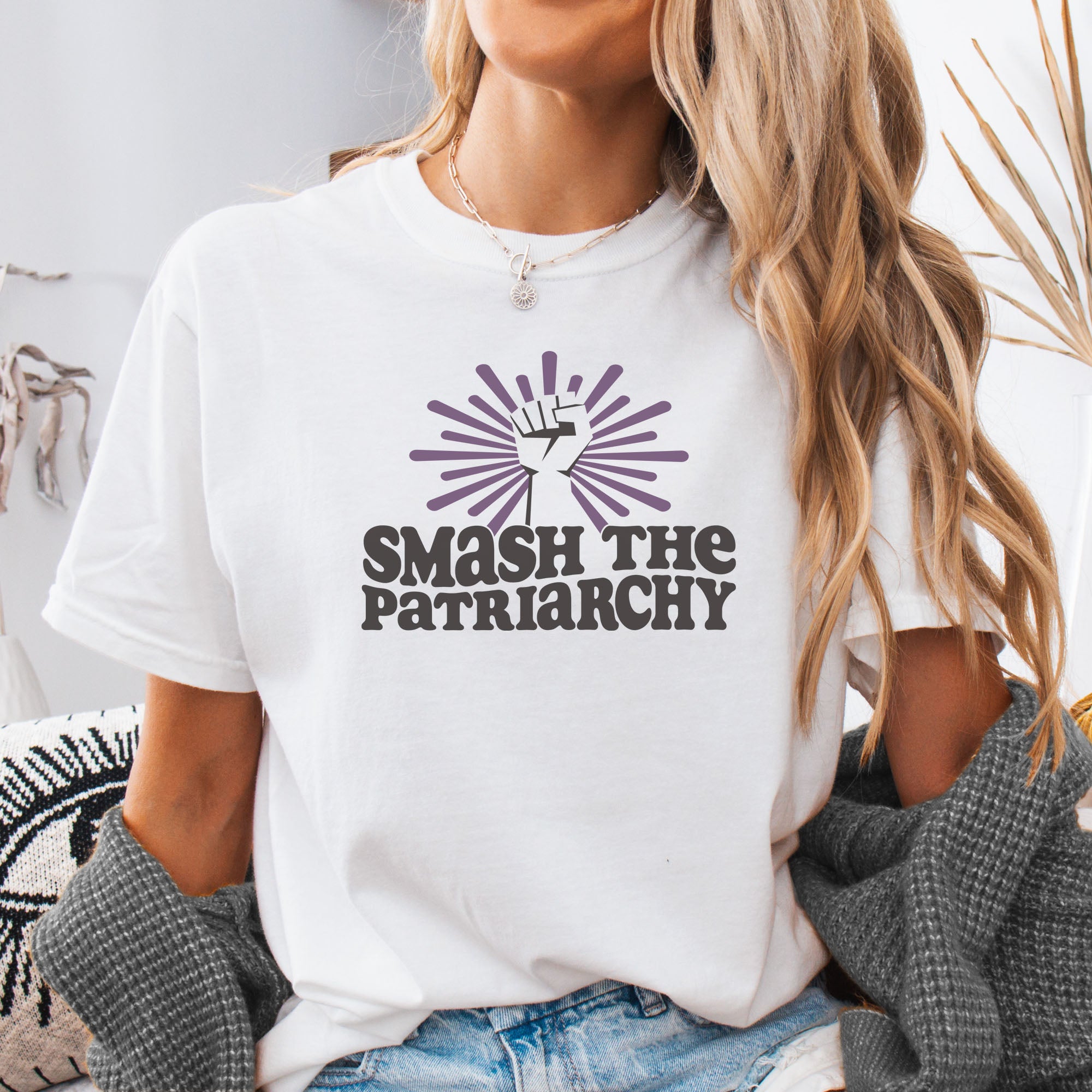 Female model wearing white t-shirt with a raised fist graphic and purple burst above the text “Smash the Patriarchy.”