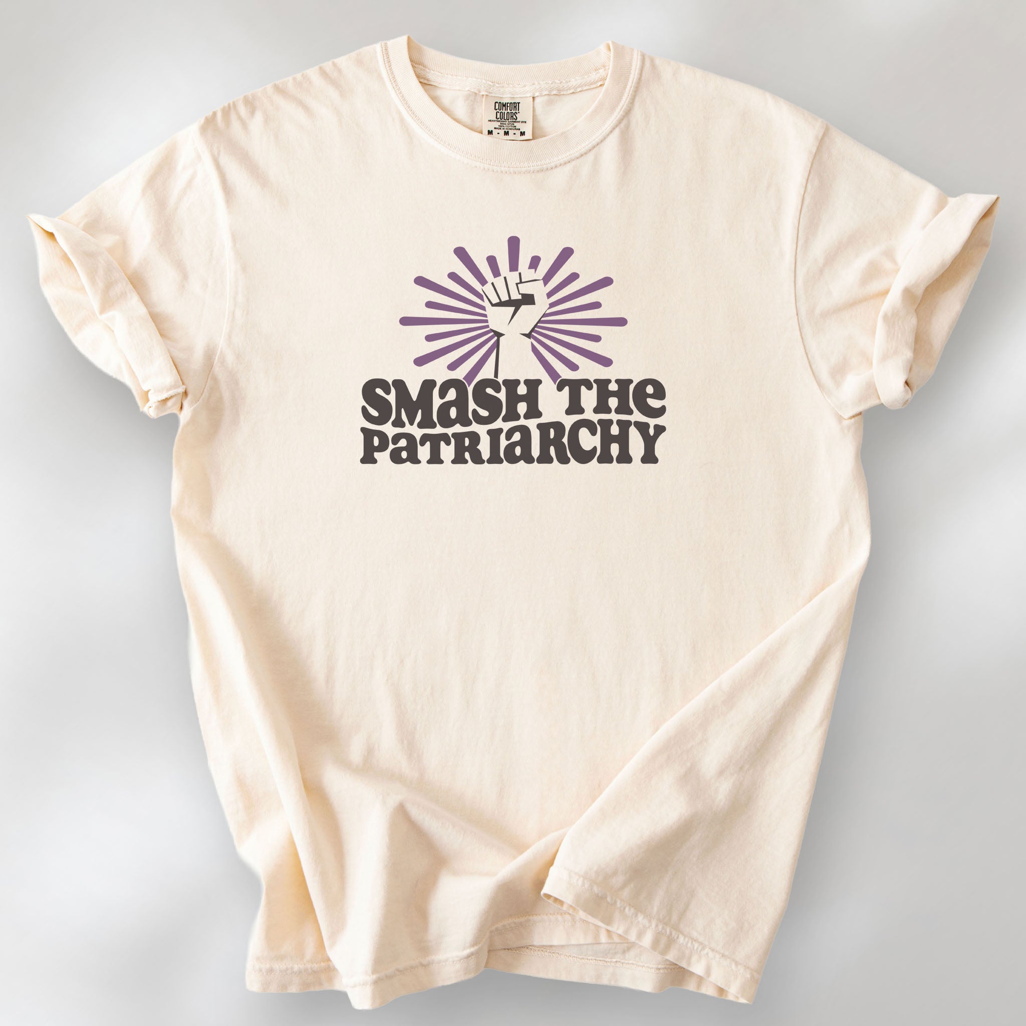 Ivory t-shirt with a raised fist graphic and purple burst above the text “Smash the Patriarchy.”