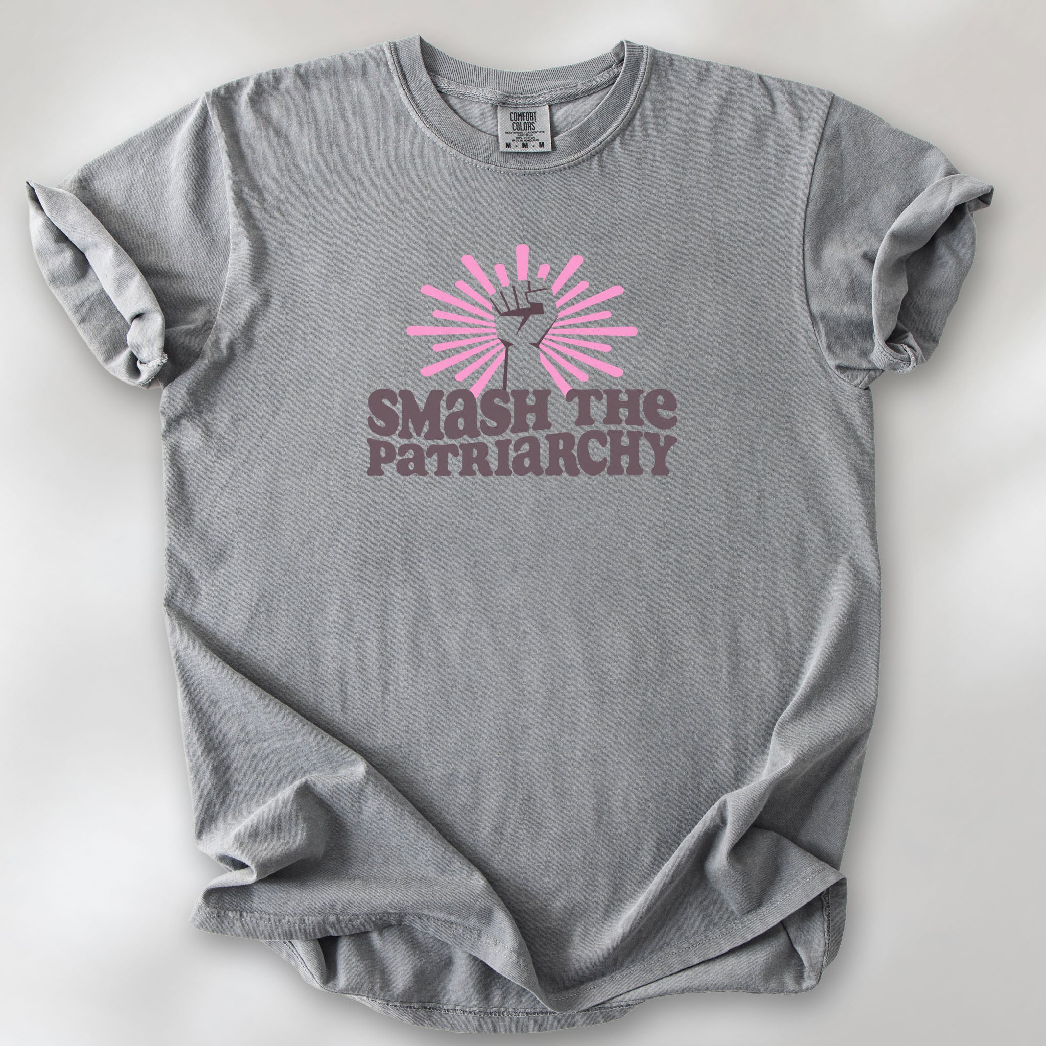 Grey t-shirt with a raised fist graphic and pink rays behind it, featuring the text “Smash the Patriarchy.”
