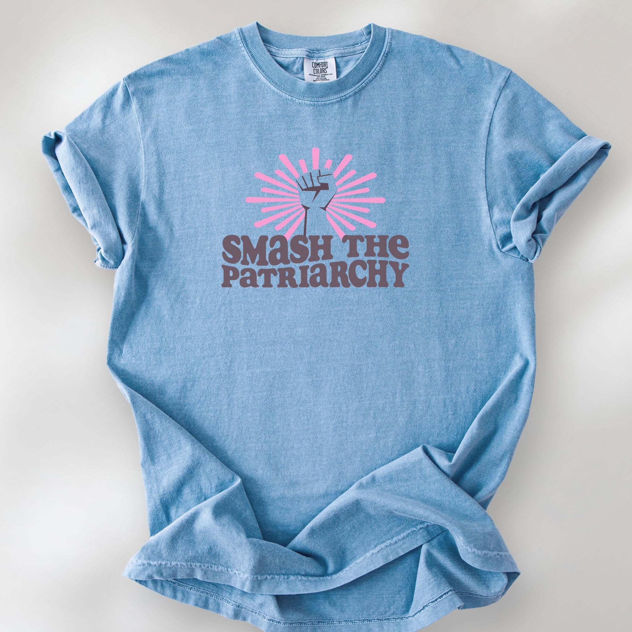 Washed denim t-shirt with a raised fist graphic and pink rays behind it, featuring the text “Smash the Patriarchy.”