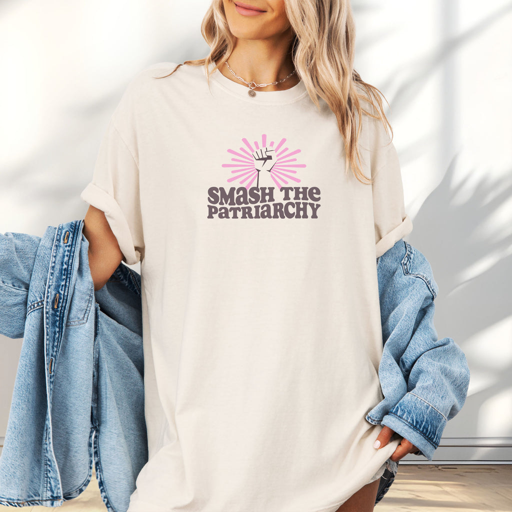 Female model wearing oversized ivory t-shirt with a raised fist graphic and pink rays behind it, featuring the text “Smash the Patriarchy.”