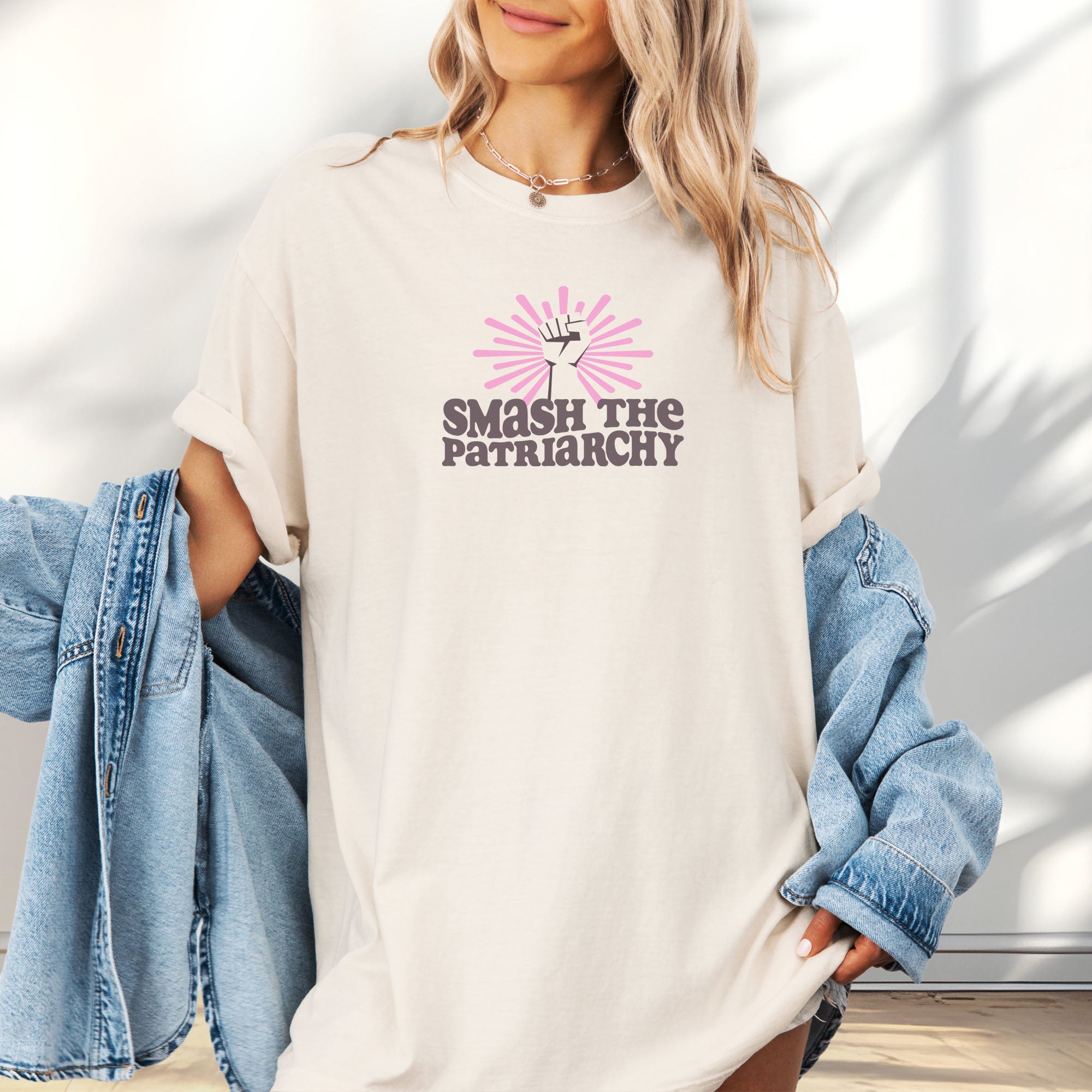 Female model wearing oversized ivory t-shirt with a raised fist graphic and pink rays behind it, featuring the text “Smash the Patriarchy.”