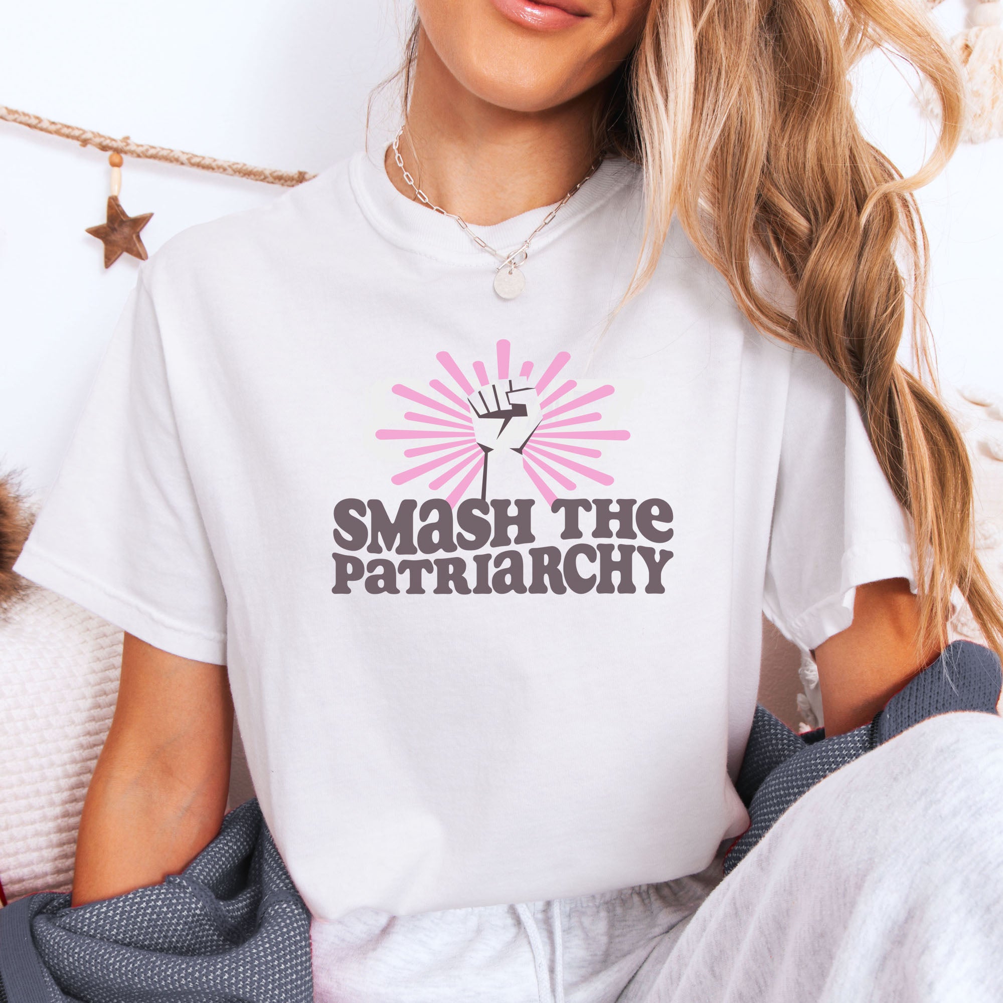 Female model wearing white t-shirt with a raised fist graphic and pink rays behind it, featuring the text “Smash the Patriarchy.”