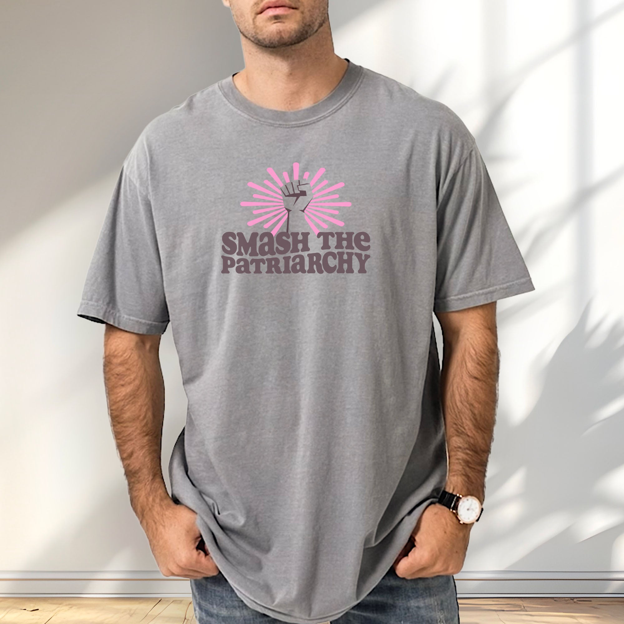 Male model wearing grey t-shirt with a raised fist graphic and pink rays behind it, featuring the text “Smash the Patriarchy.”