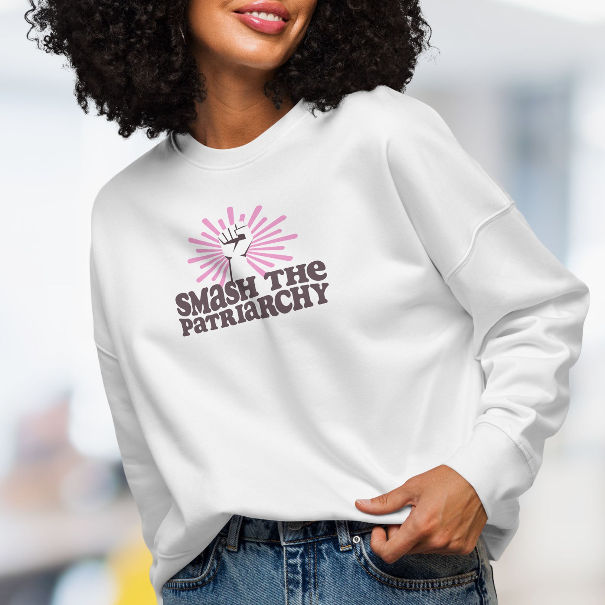 Front view of female model wearing white sweatshirt with a raised fist graphic and pink rays behind it, featuring the text “Smash the Patriarchy.”