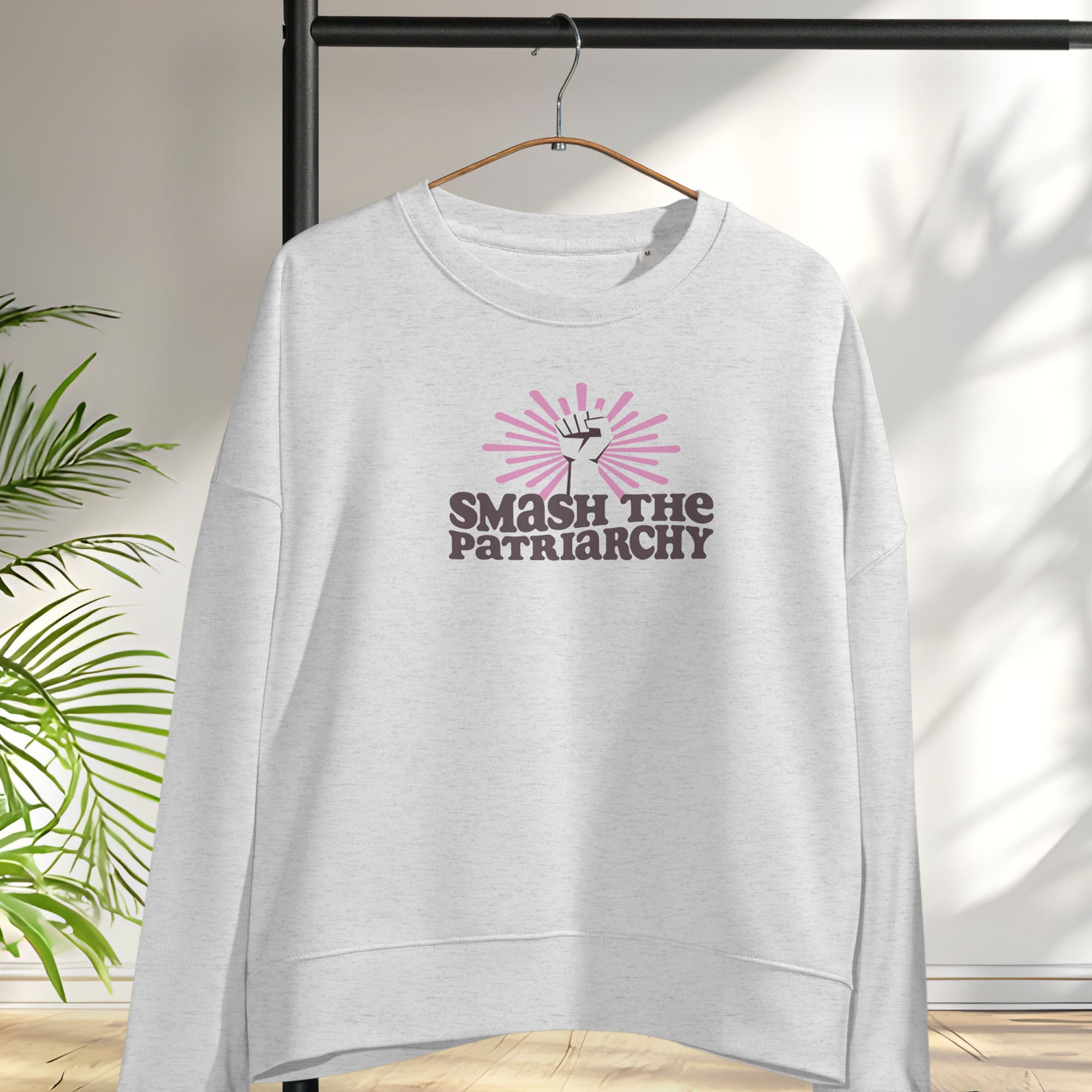 Grey sweatshirt with a raised fist graphic and pink rays behind it, featuring the text “Smash the Patriarchy.” Hanging on a hanger.