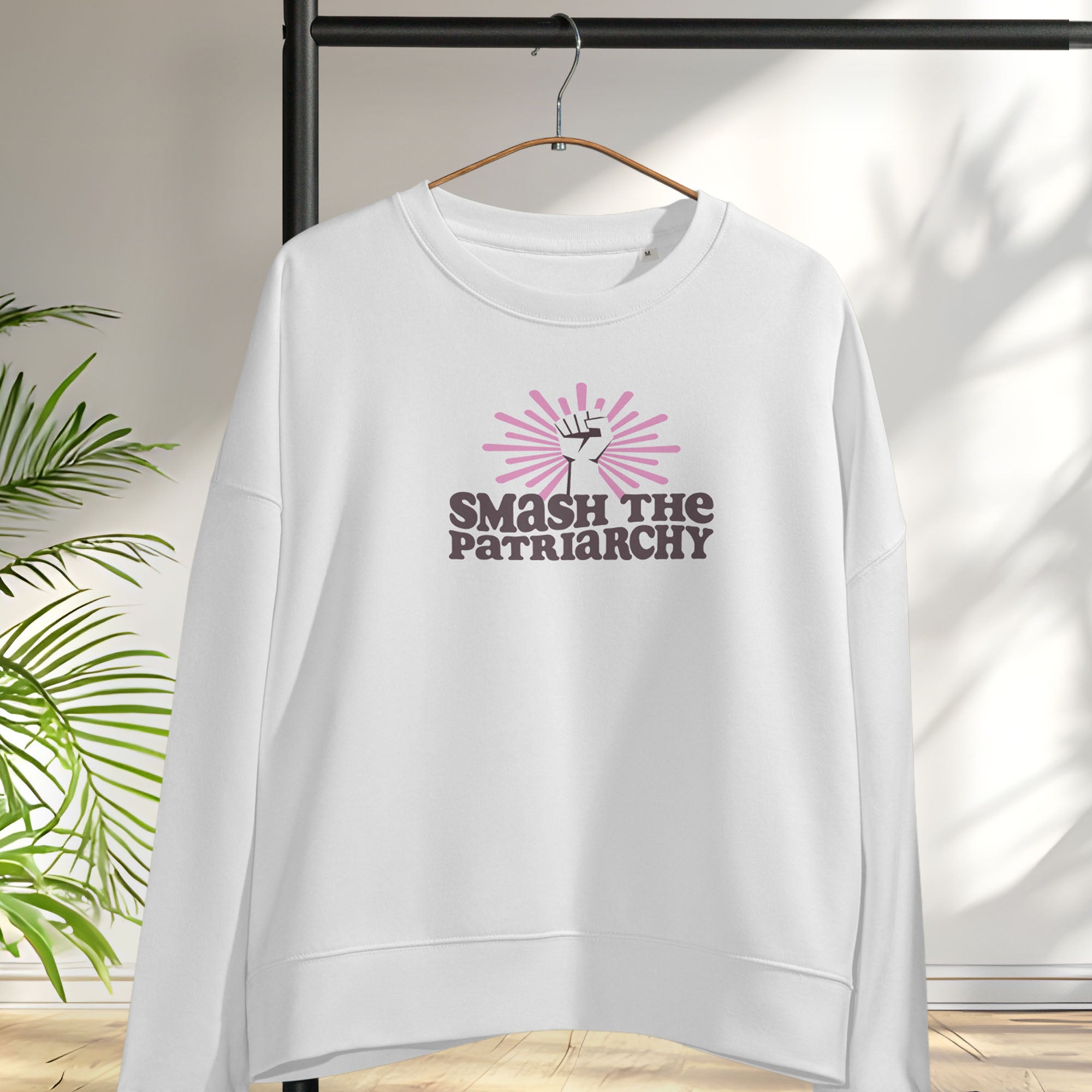White sweatshirt with a raised fist graphic and pink rays behind it, featuring the text “Smash the Patriarchy.” Hanging on a hanger.