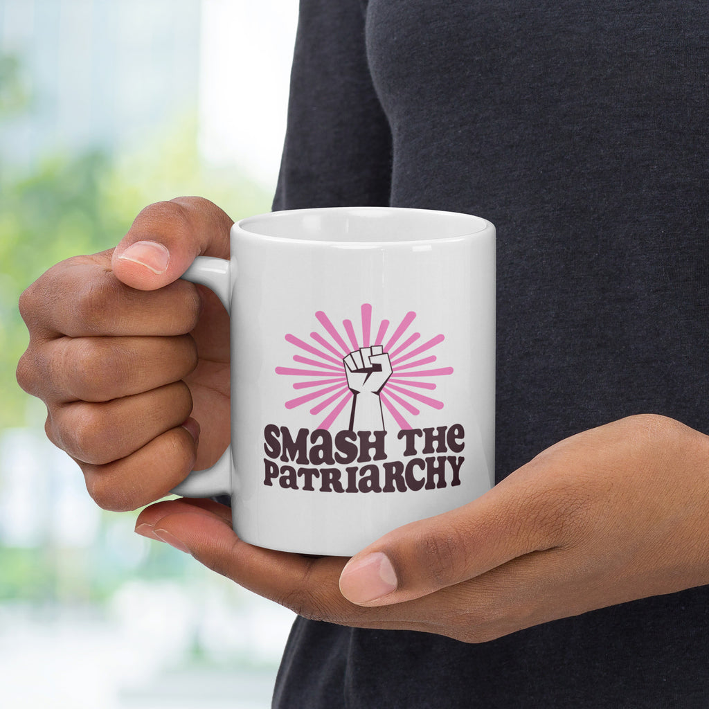 Person holding coffee mug that says "smash the patriarchy" below a raised fist centered in front of pink rays, with handle on left.