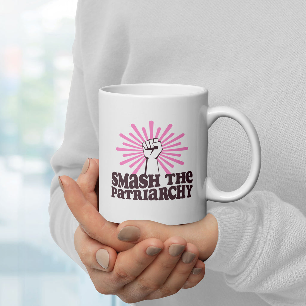 Person holding coffee mug that says "smash the patriarchy" below a raised fist centered in front of pink rays, with handle on right.