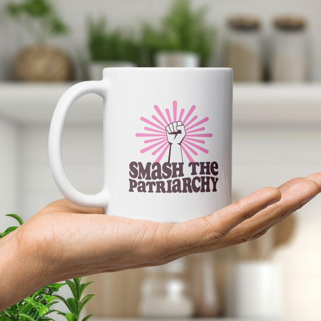 Coffee mug that says "smash the patriarchy" below a raised fist centered in front of pink rays, held in the palm of a hand.
