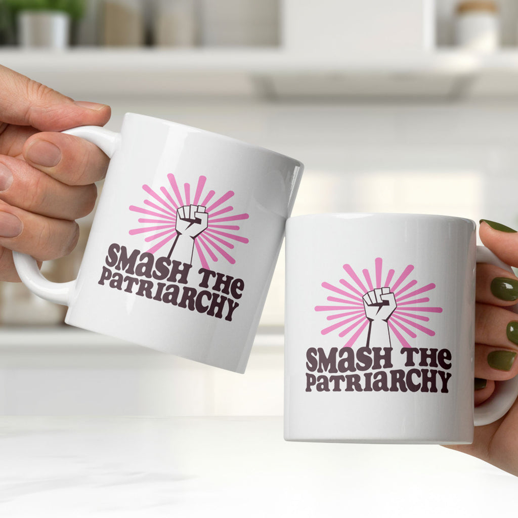 Two people clinking coffee mugs that says "smash the patriarchy" below a raised fist centered in front of pink rays.