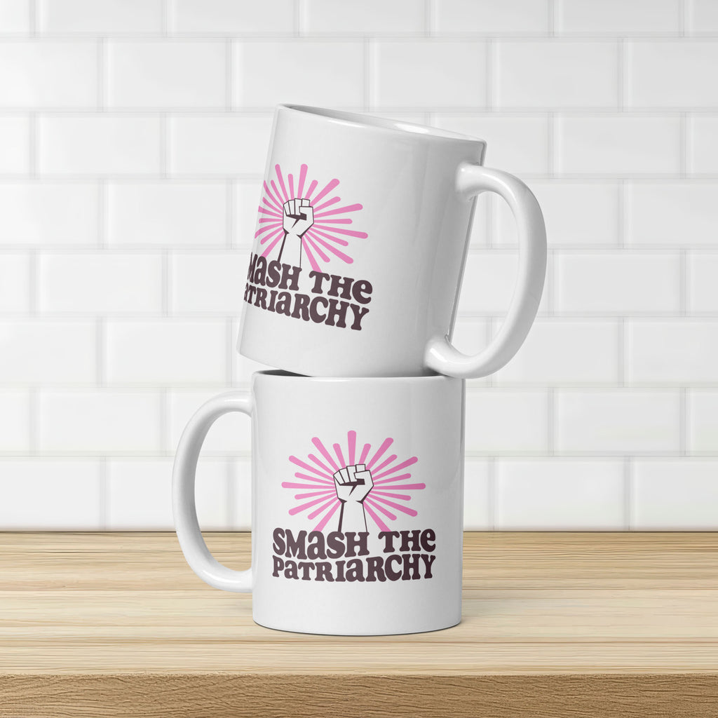 Two stacked coffee mugs that say "smash the patriarchy" below a raised fist centered in front of pink rays, showing same design on both sides of mug.