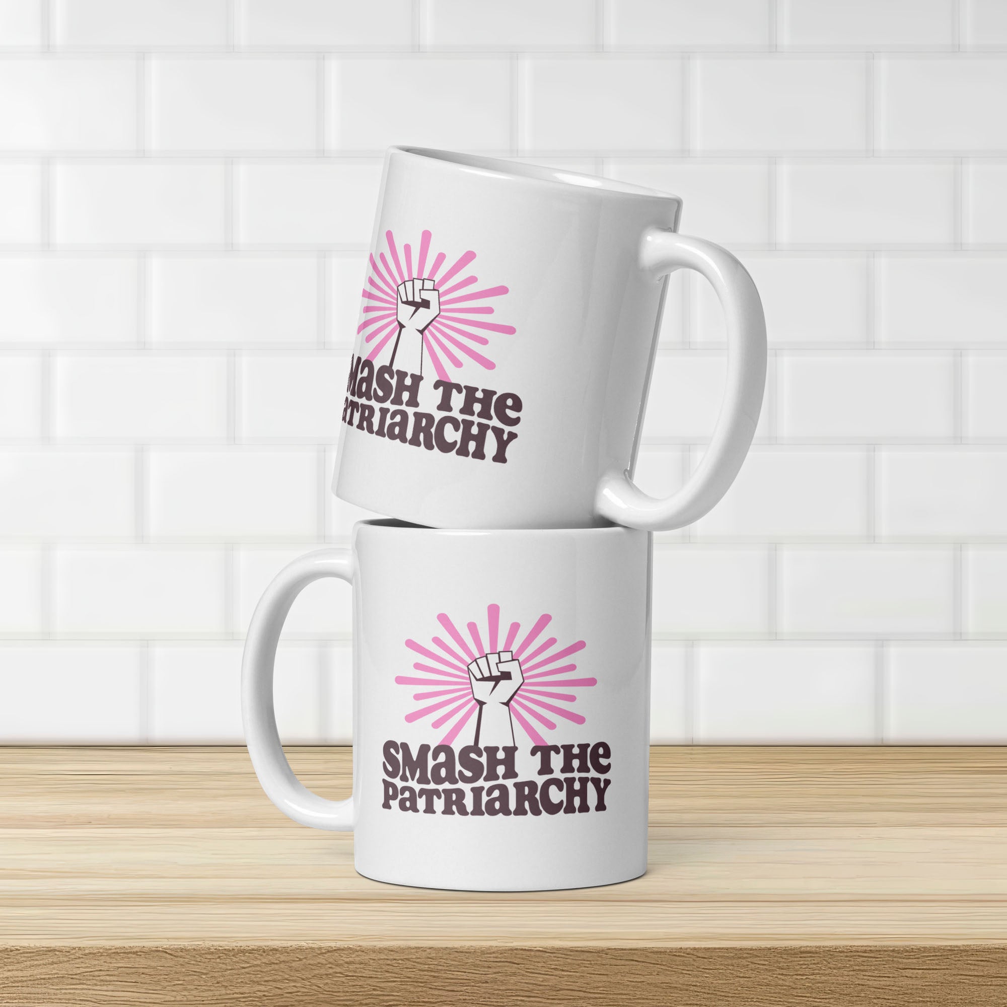 Two stacked coffee mugs that say "smash the patriarchy" below a raised fist centered in front of pink rays, showing same design on both sides of mug.