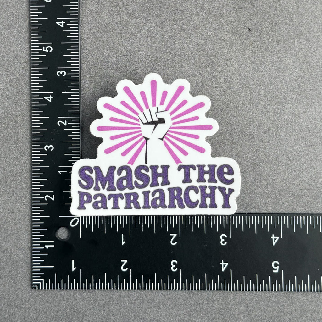 Die-cut vinyl sticker featuring a raised fist with a pink sunburst above the words "Smash the Patriarchy" in bold purple lettering, with ruler to show the approximate size of 3 inches by 3 inches.