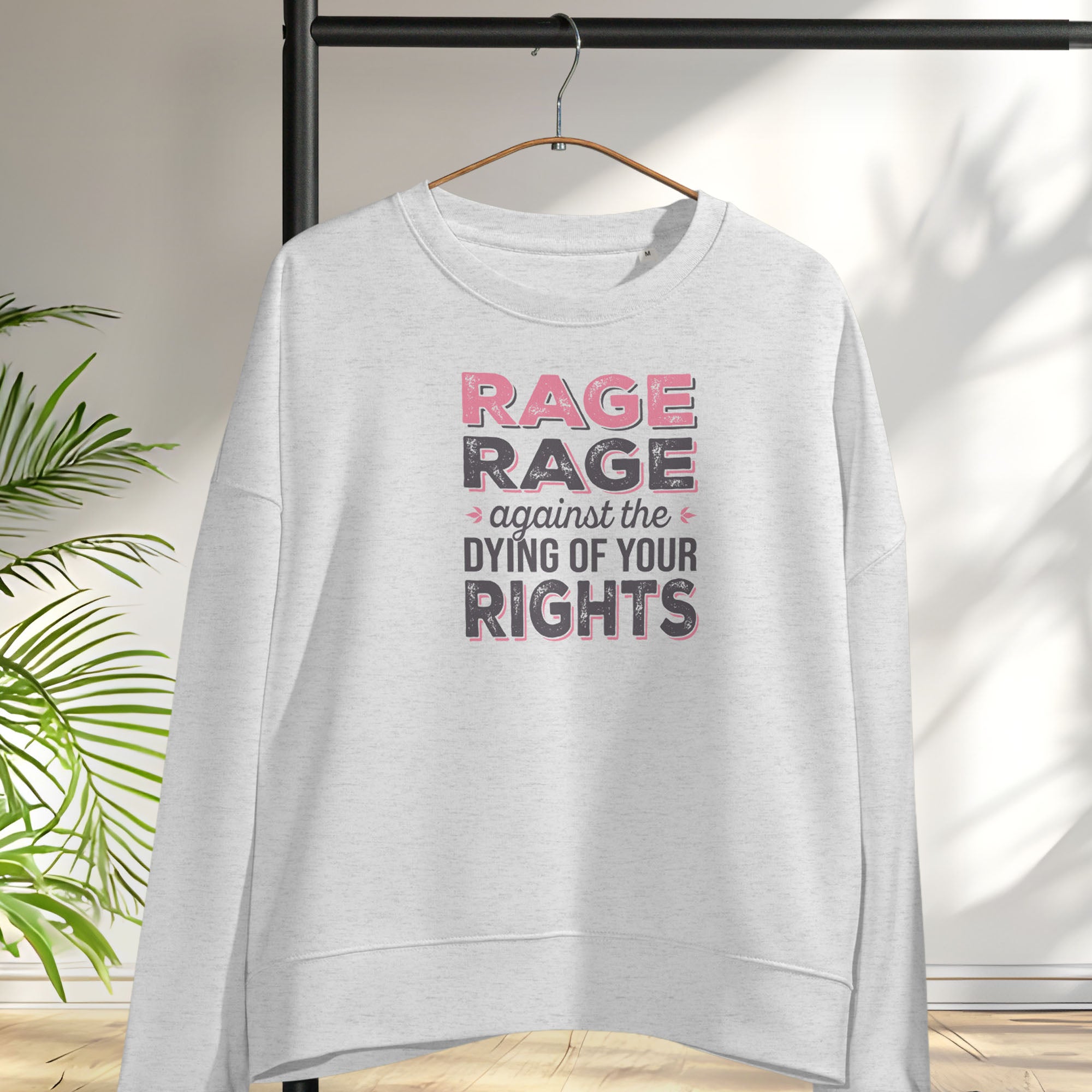 Grey women's protest sweatshirt with bold pink and grey text reading “rage rage against the dying of your rights” – feminist anti-trump activist shirt, hanging on a hanger.