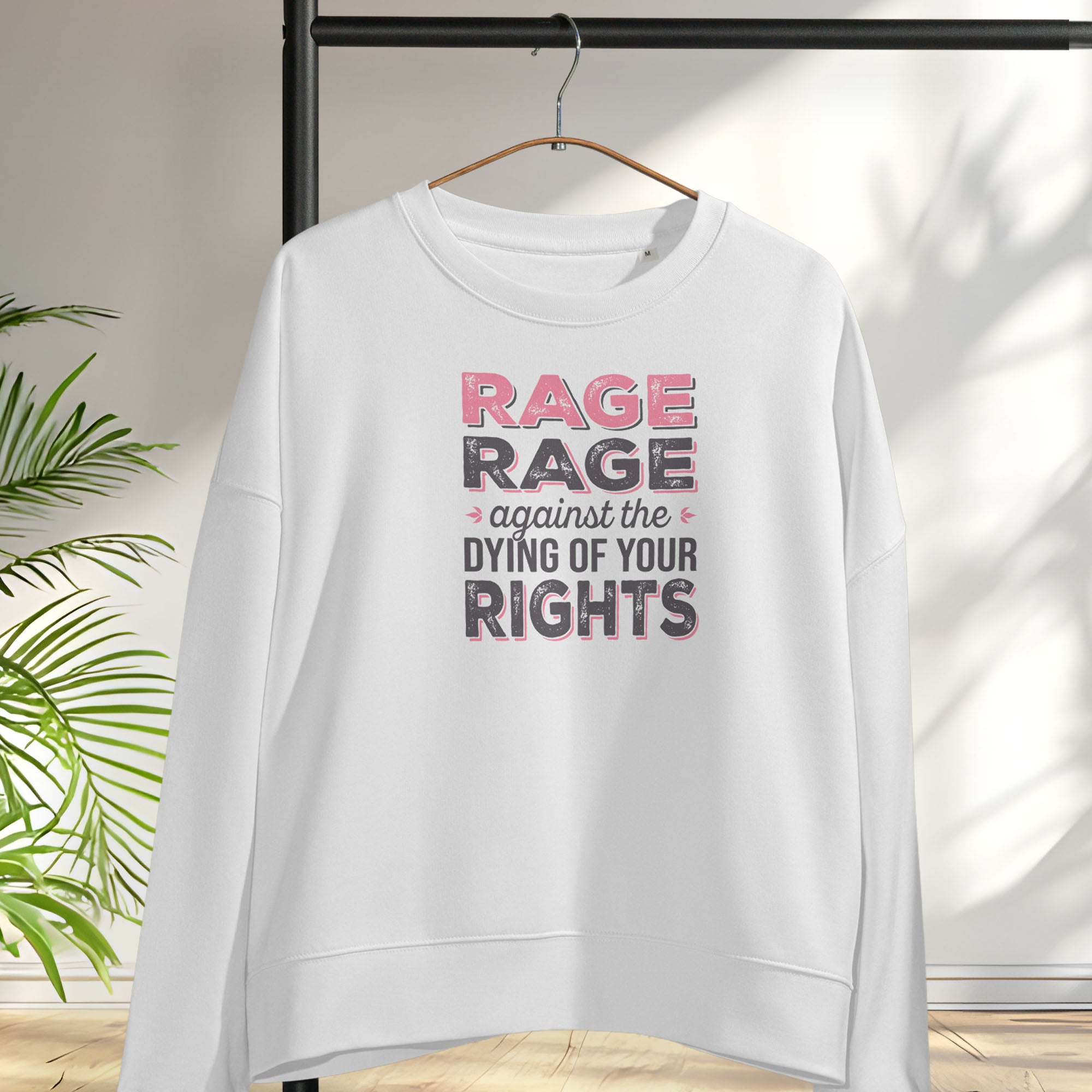 White women's protest sweatshirt with bold pink and grey text reading “rage rage against the dying of your rights” – feminist anti-trump activist shirt, hanging on a hanger.