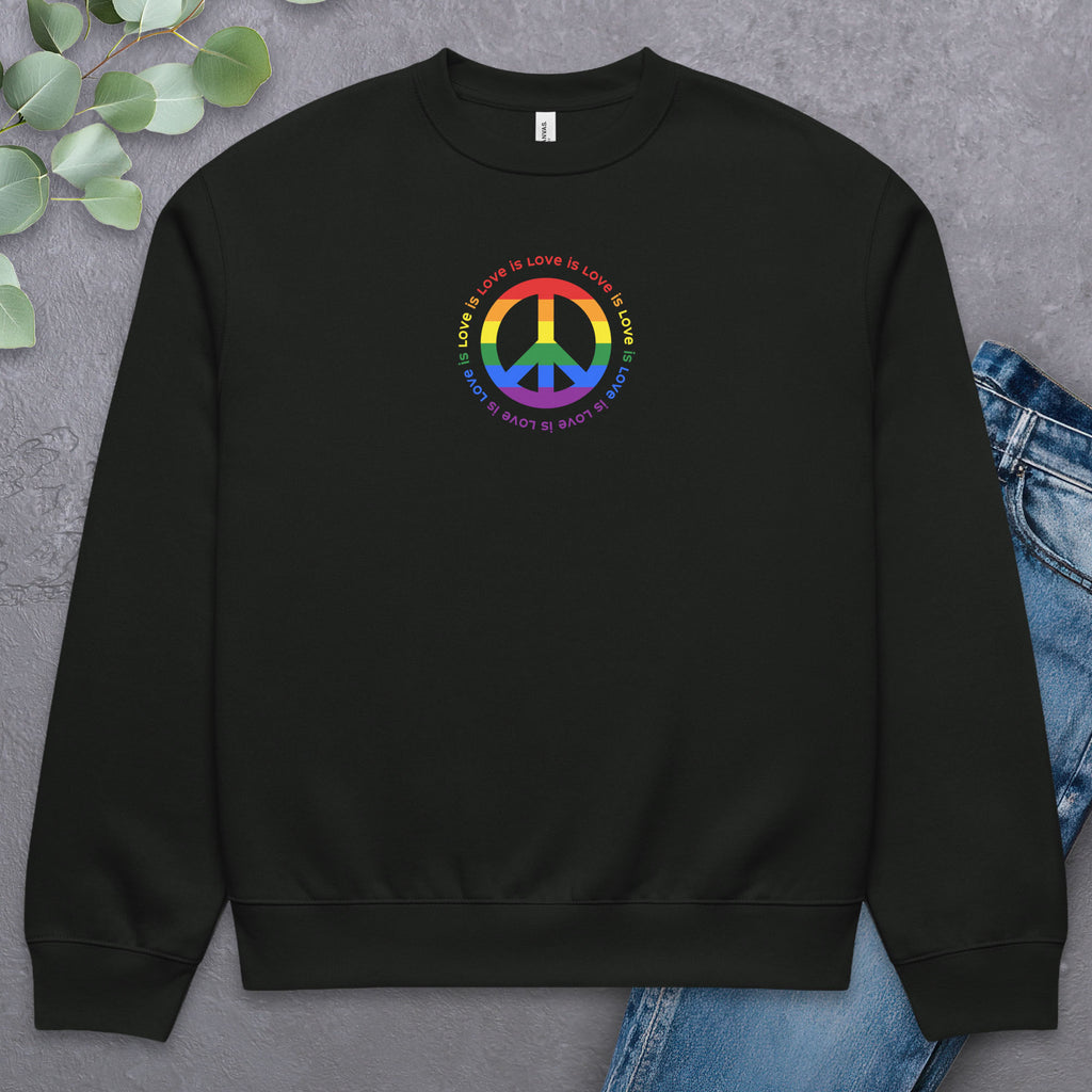 Black sweatshirt with LGBTQ pride graphic — a peace sign in the LGBTQ flag colors, with the text "love is love" repeated in a circle around the peace sign. Laying flat with a pair of jeans.