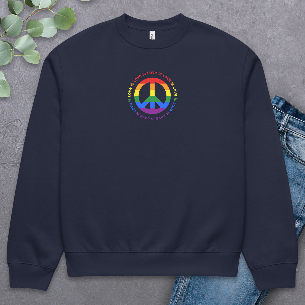 Navy sweatshirt with LGBTQ pride graphic — a peace sign in the LGBTQ flag colors, with the text "love is love" repeated in a circle around the peace sign. Laying flat with a pair of jeans.