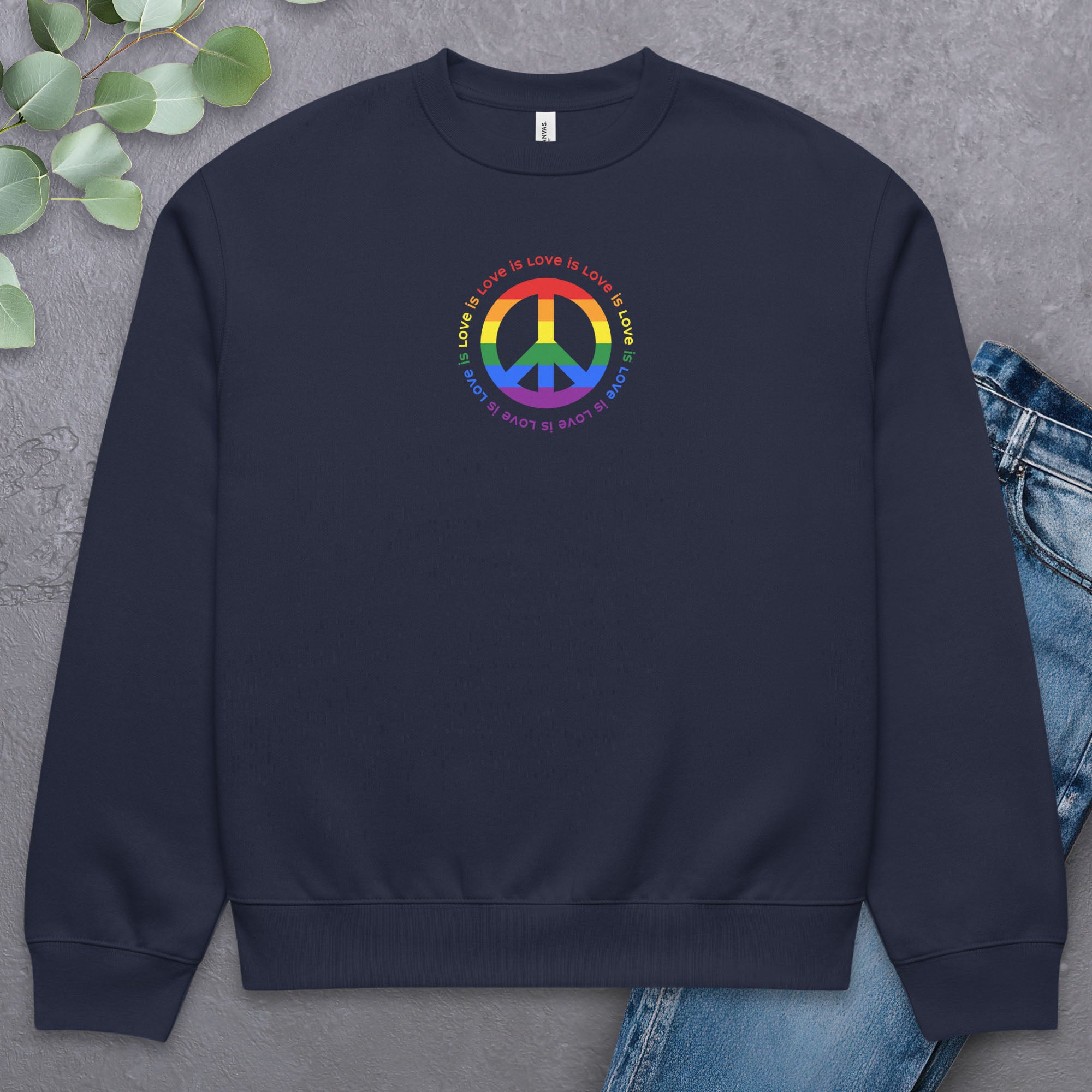 Navy sweatshirt with LGBTQ pride graphic — a peace sign in the LGBTQ flag colors, with the text "love is love" repeated in a circle around the peace sign. Laying flat with a pair of jeans.