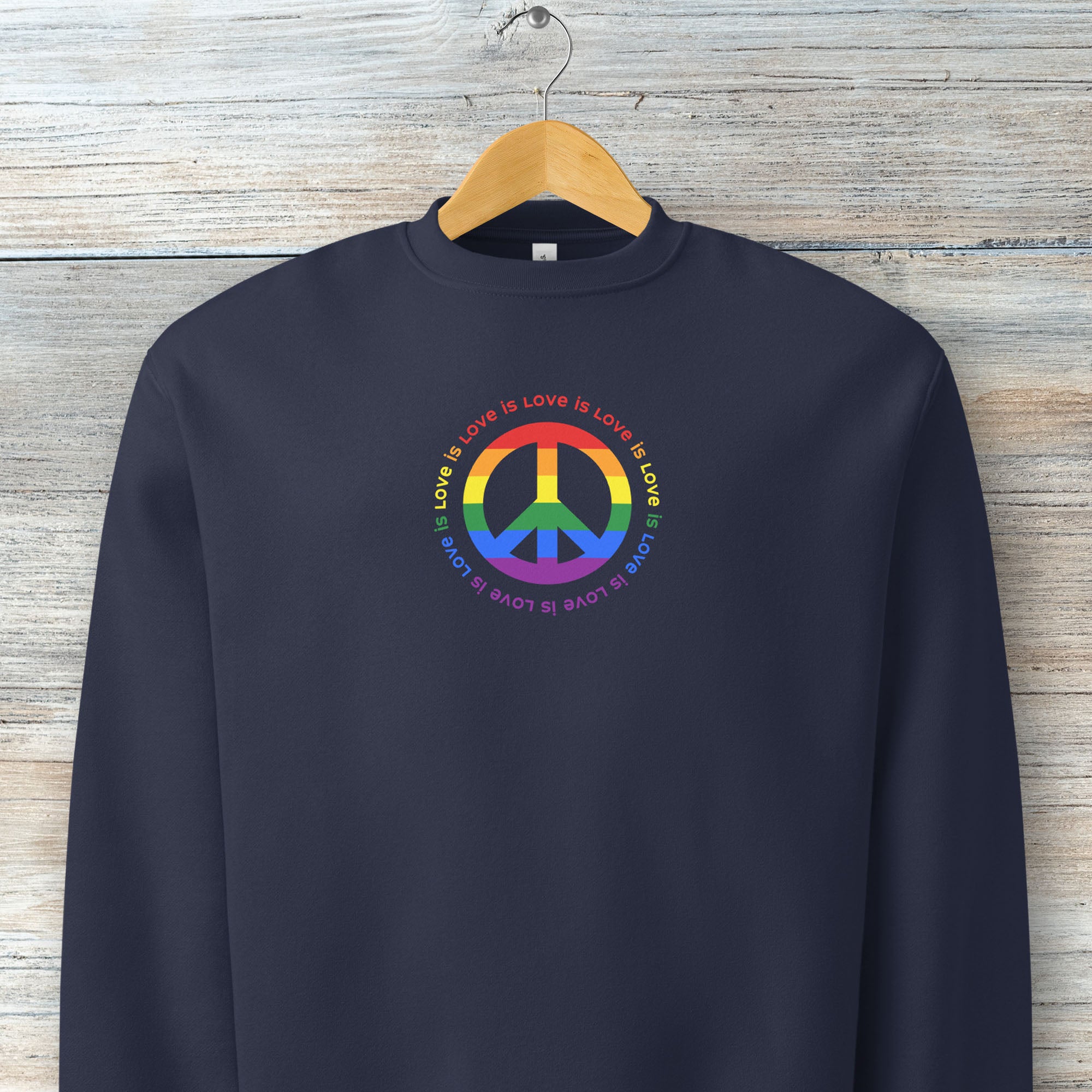 Navy sweatshirt with LGBTQ pride graphic — a peace sign in the LGBTQ flag colors, with the text "love is love" repeated in a circle around the peace sign. Hanging on a hanger.