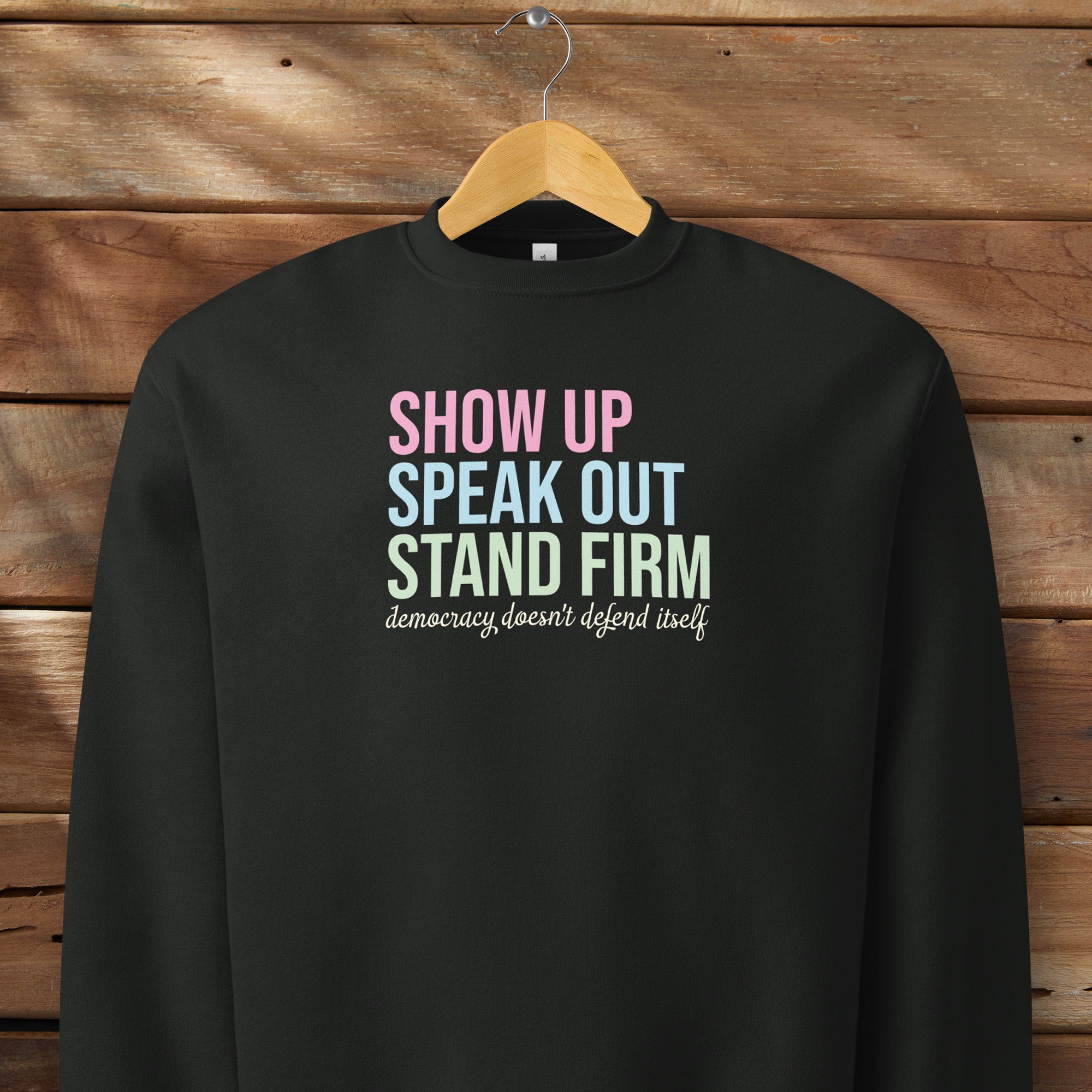 Black unisex crewneck sweatshirt with bright bold text that reads "show up, speak out, stand firm" then smaller script text that reads "democracy doesn't defend itself", on a hanger.