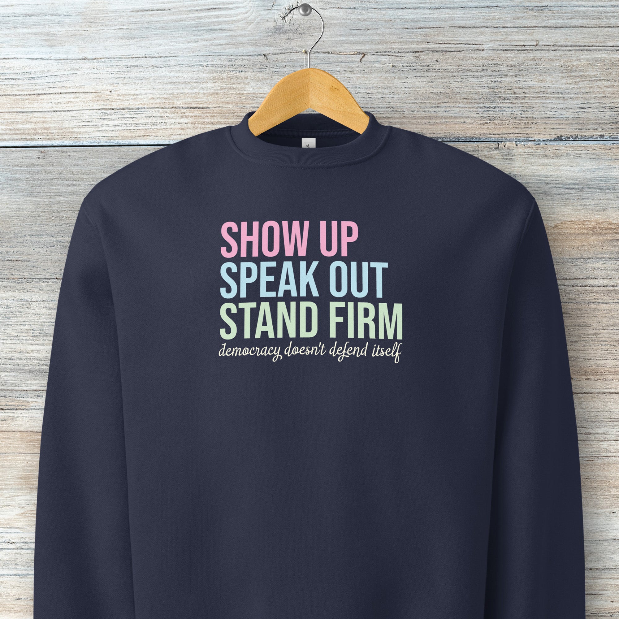 Navy unisex crewneck sweatshirt with bright bold text that reads "show up, speak out, stand firm" then smaller script text that reads "democracy doesn't defend itself", on a hanger.