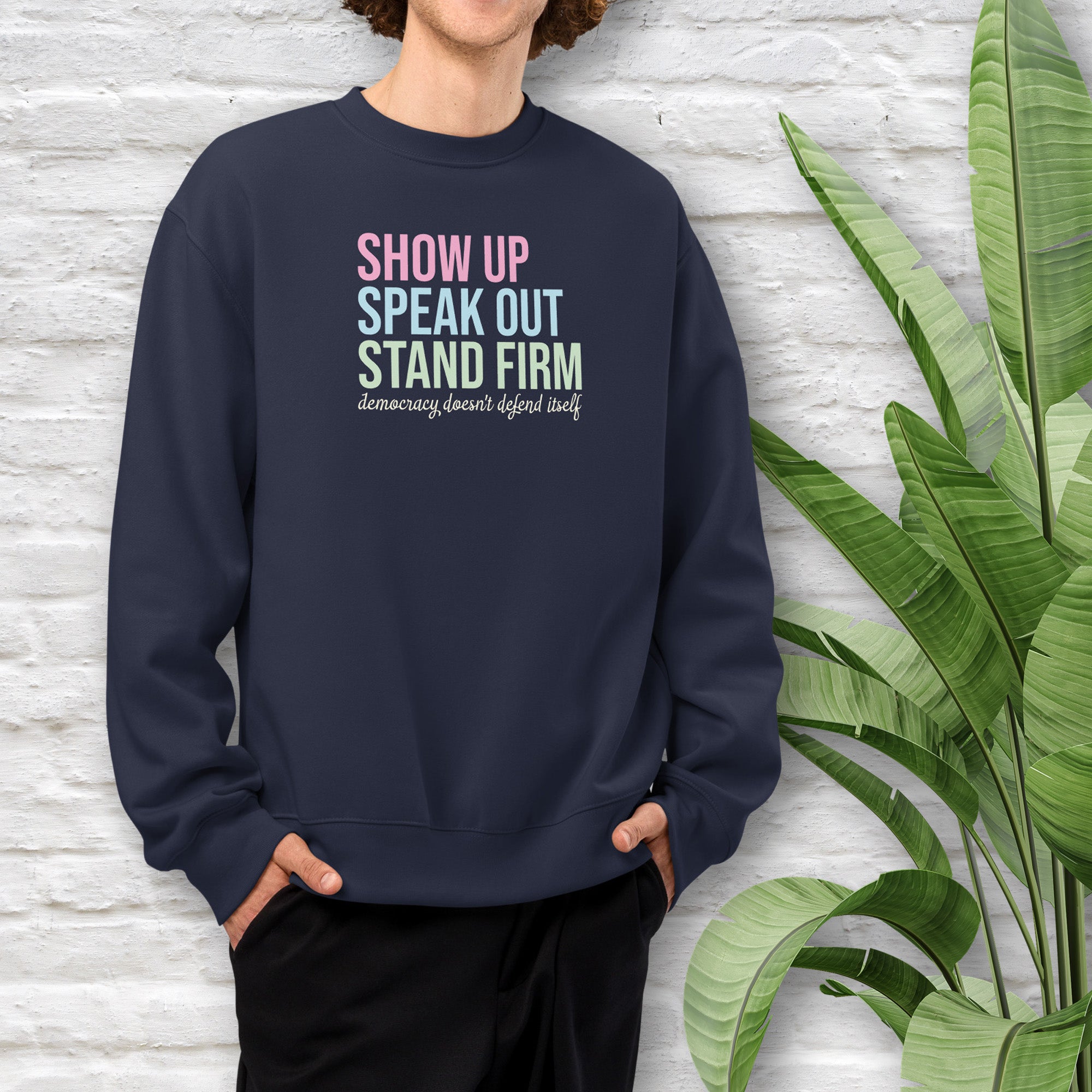 Male model wearing navy unisex crewneck sweatshirt with bright bold text that reads "show up, speak out, stand firm" then smaller script text that reads "democracy doesn't defend itself".