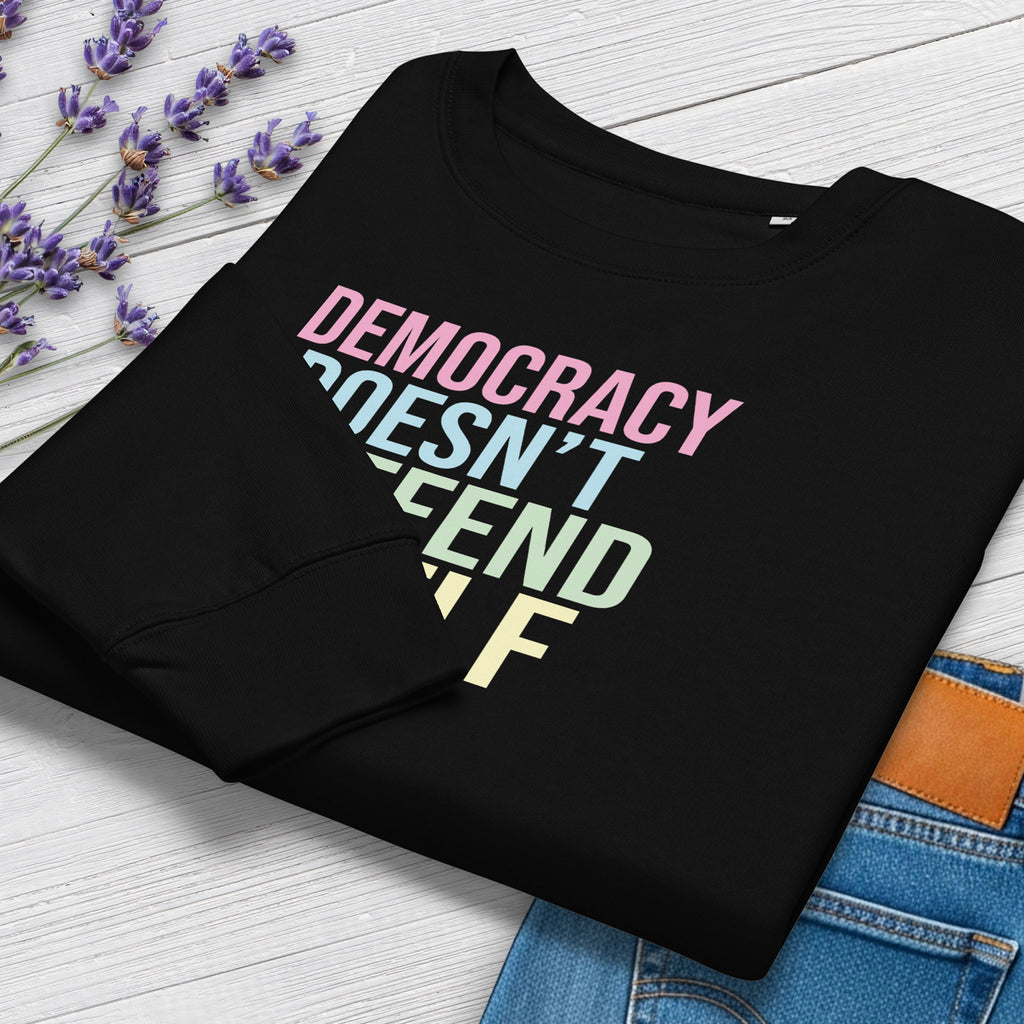 Folded women's black sweatshirt with bright pink, blue, green, and yellow bold text that says "democracy doesn't defend itself." Styled with jeans and flowers.