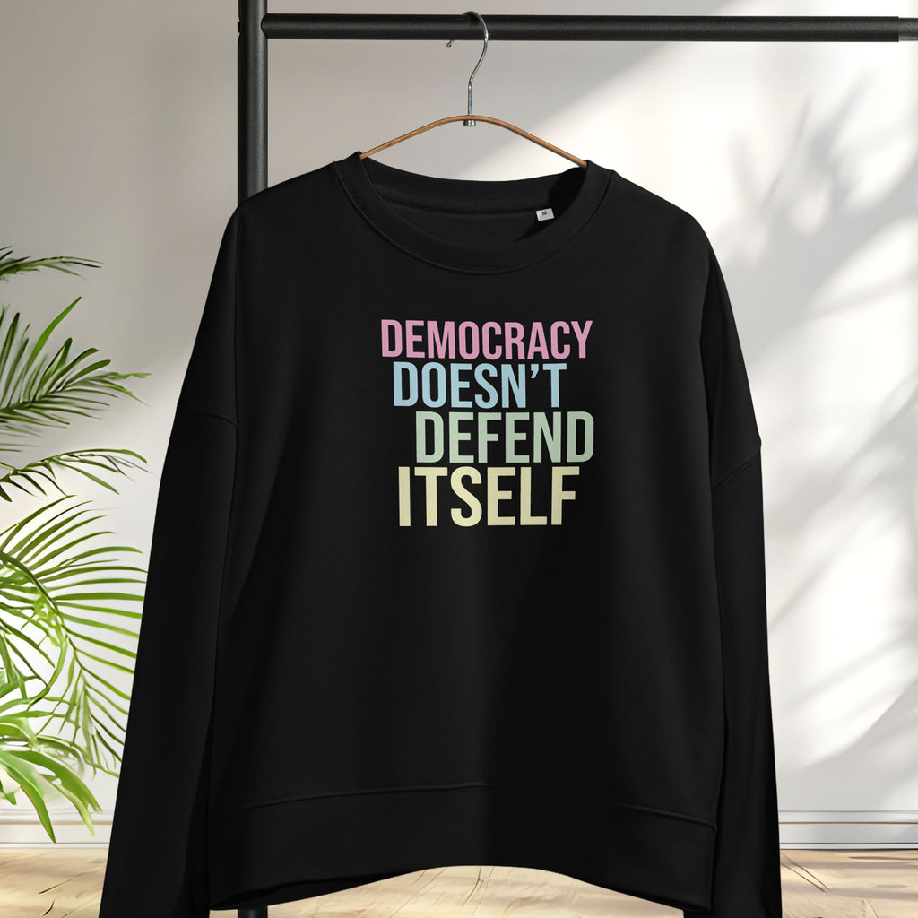 Women's black sweatshirt with bright pink, blue, green, and yellow bold text that says "democracy doesn't defend itself." Hanging on a hanger.