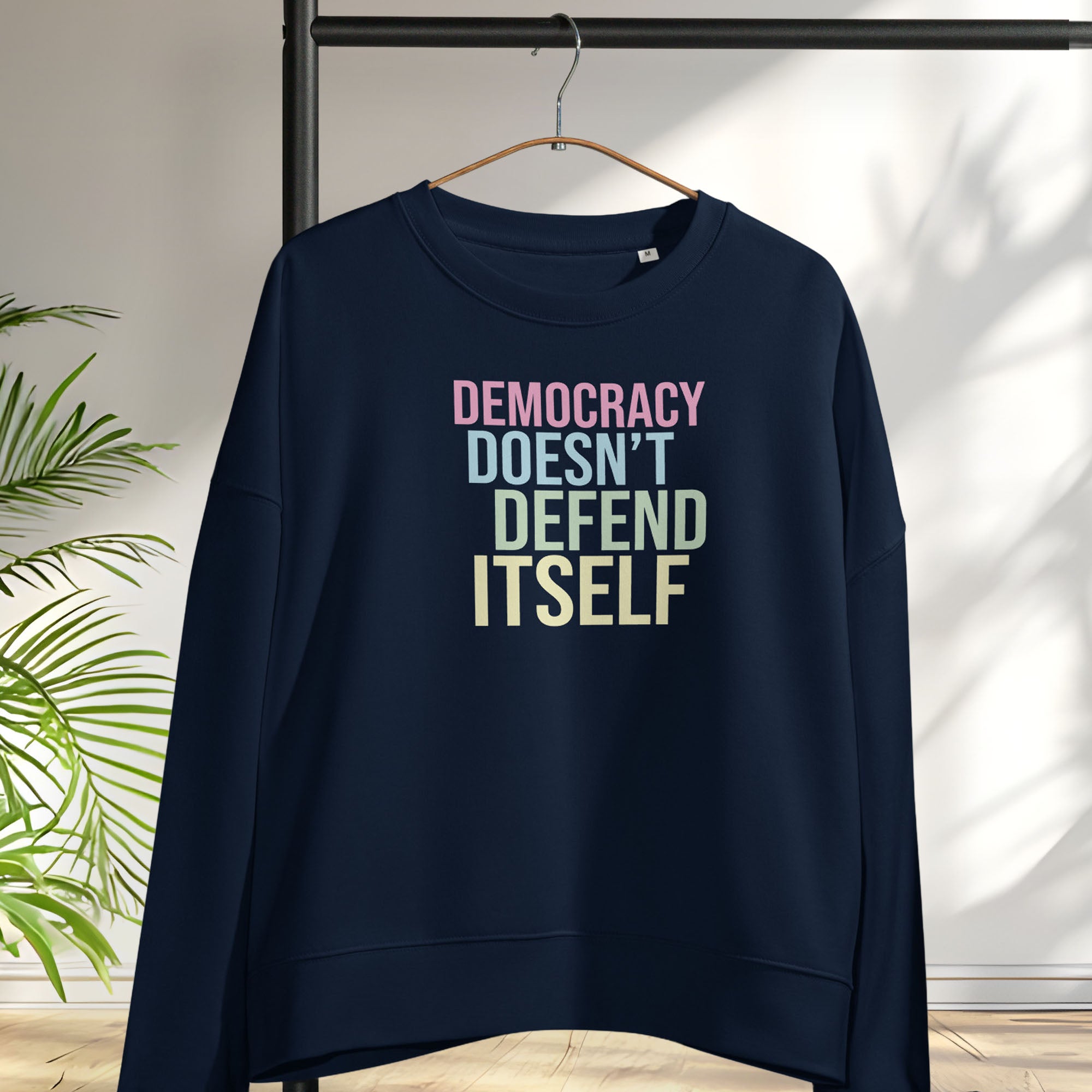 Women's navy sweatshirt with bright pink, blue, green, and yellow bold text that says "democracy doesn't defend itself." Hanging on a hanger.