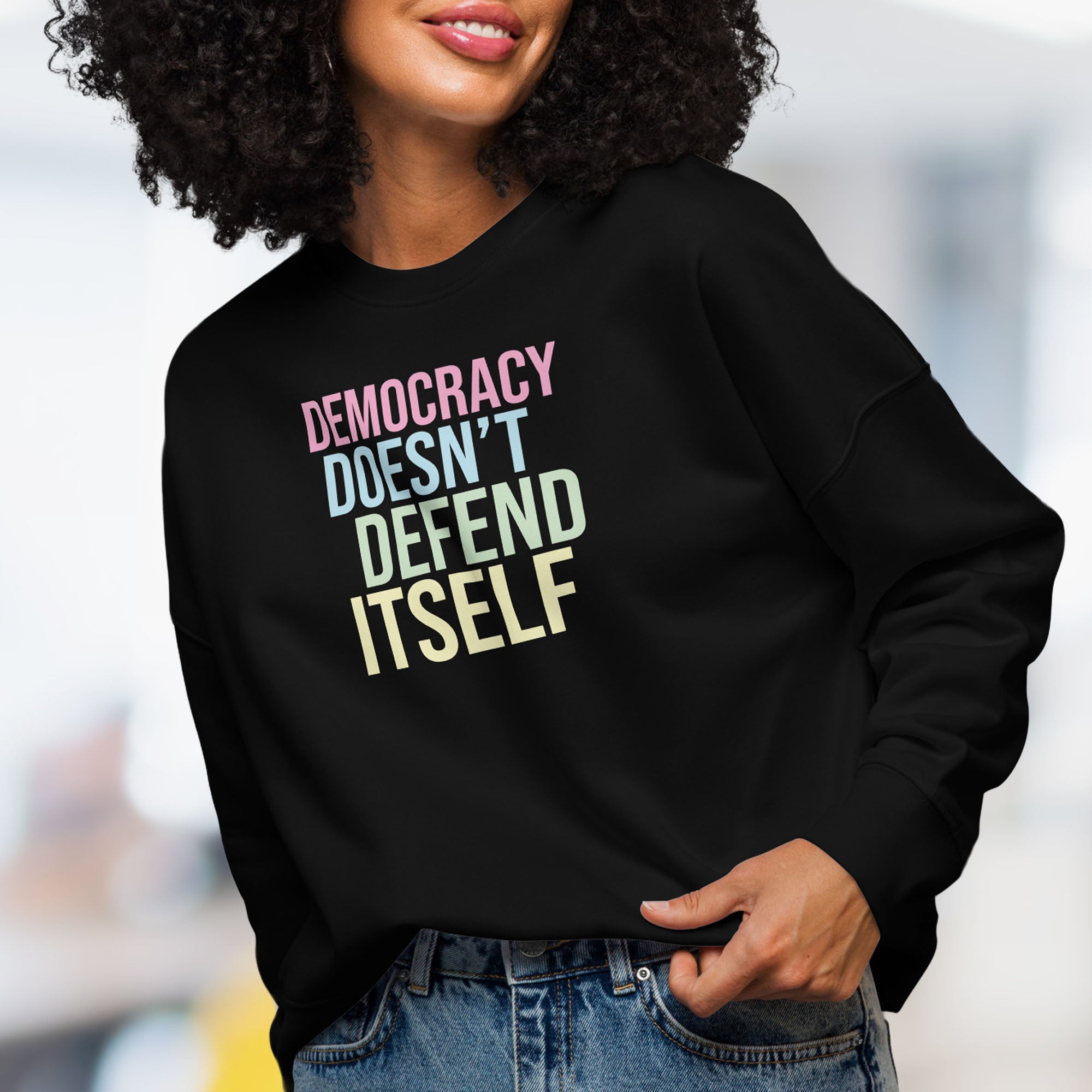 Female model wearing women's black sweatshirt with bright pink, blue, green, and yellow bold text that says "democracy doesn't defend itself."