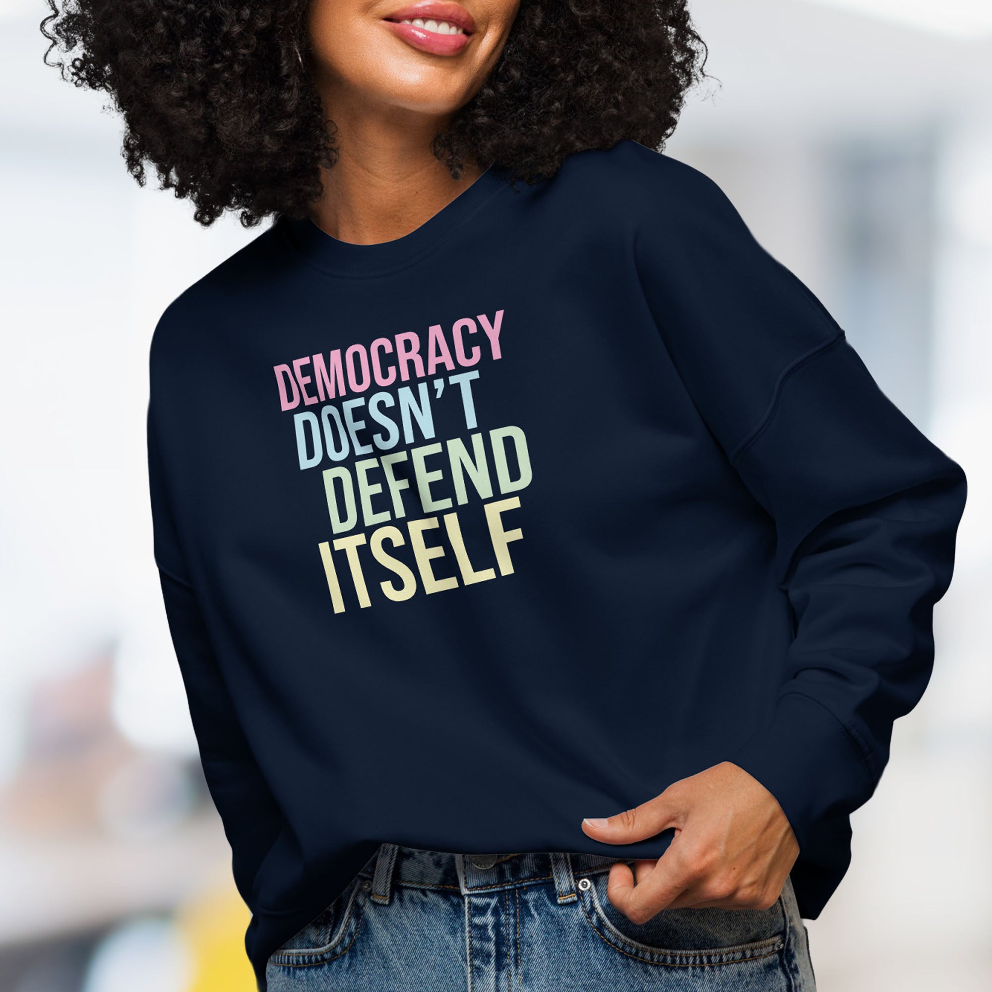Female model wearing women's navy sweatshirt with bright pink, blue, green, and yellow bold text that says "democracy doesn't defend itself."