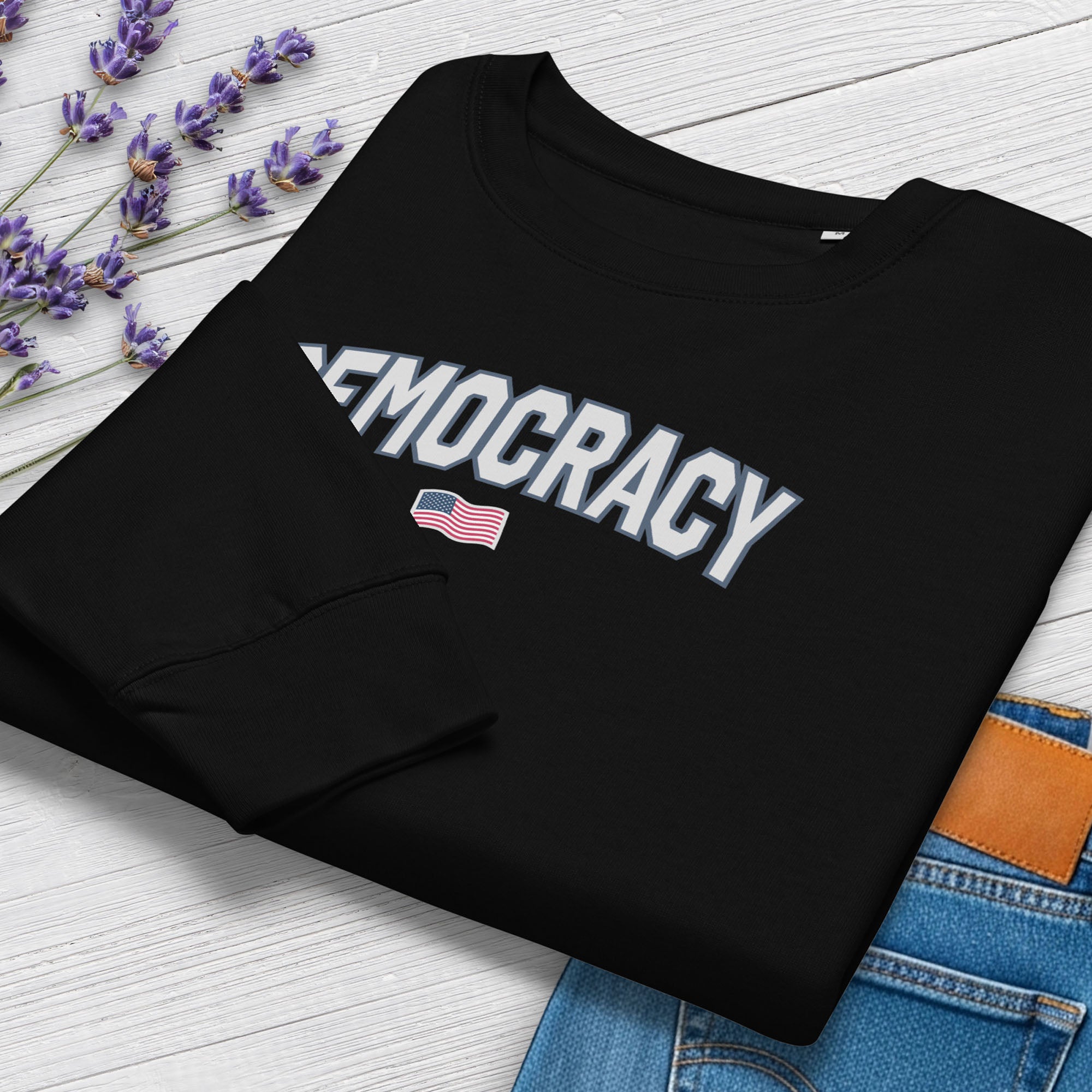 Folded women's black sweatshirt with the word “Democracy” in bold grey letters and a small American flag underneath, symbolizing U.S. pride in democracy over authoritarianism, tyranny, dictators, handing on a hanger.