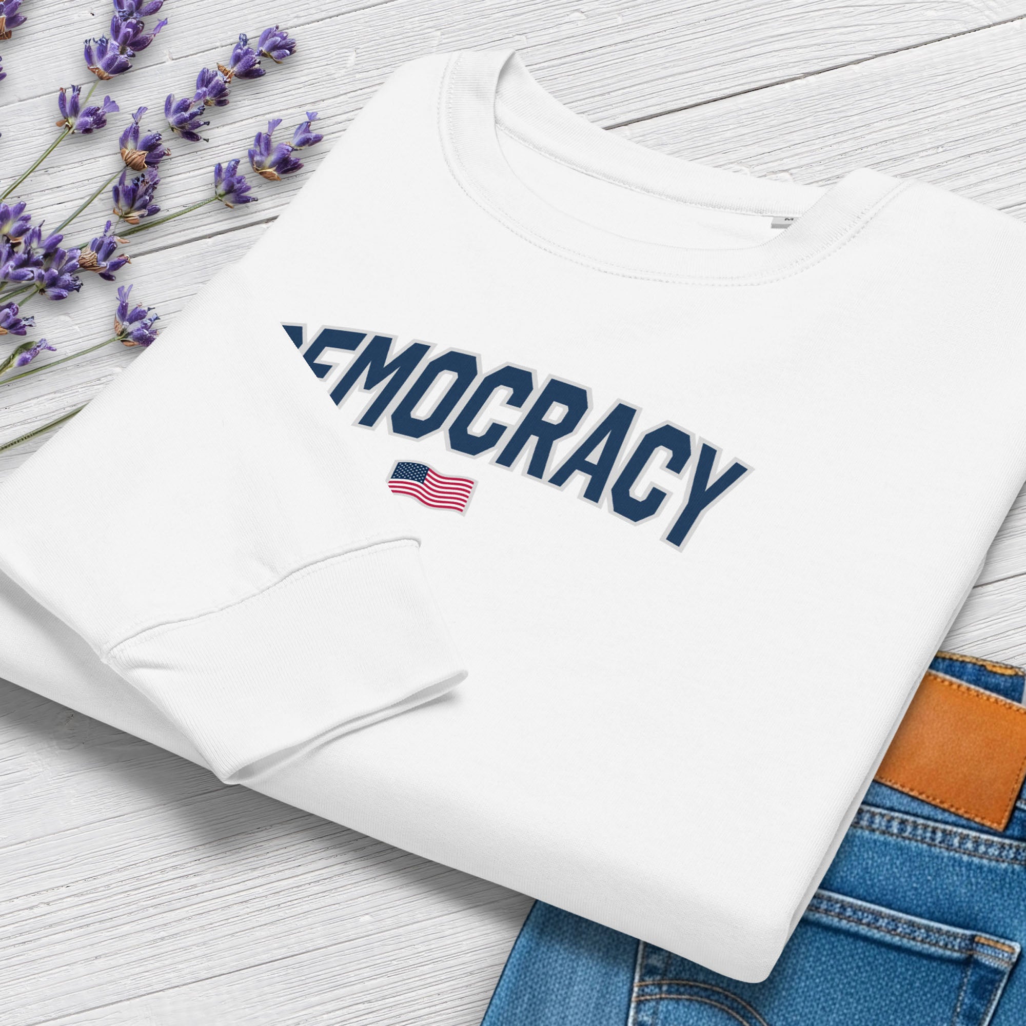 Folded women's white sweatshirt with the word “Democracy” in bold navy letters and a small American flag underneath, symbolizing U.S. pride in democracy over authoritarianism, tyranny, dictators, handing on a hanger.