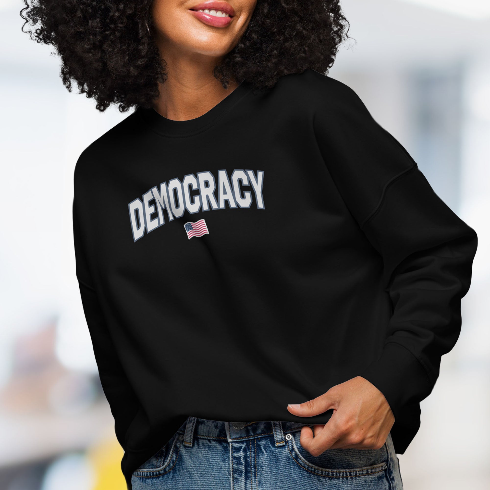 Front view of female model wearing women's black sweatshirt with the word “Democracy” in bold grey letters and a small American flag underneath, symbolizing U.S. pride in democracy over authoritarianism, tyranny, dictators.