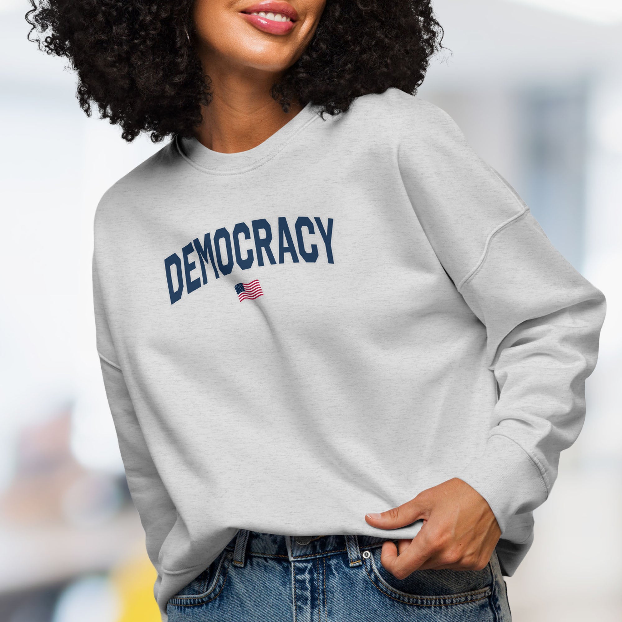 Front view of female model wearing women's grey sweatshirt with the word “Democracy” in bold navy letters and a small American flag underneath, symbolizing U.S. pride in democracy over authoritarianism, tyranny, dictators.