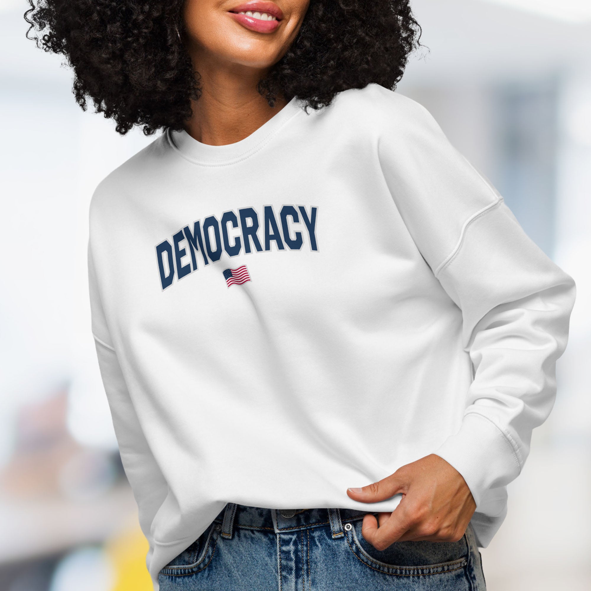 Front view of female model wearing women's white sweatshirt with the word “Democracy” in bold navy letters and a small American flag underneath, symbolizing U.S. pride in democracy over authoritarianism, tyranny, dictators.