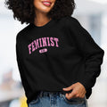 Front view of female model wearing black sweatshirt with pink text that reads “Feminist A.F.” in a collegiate style.