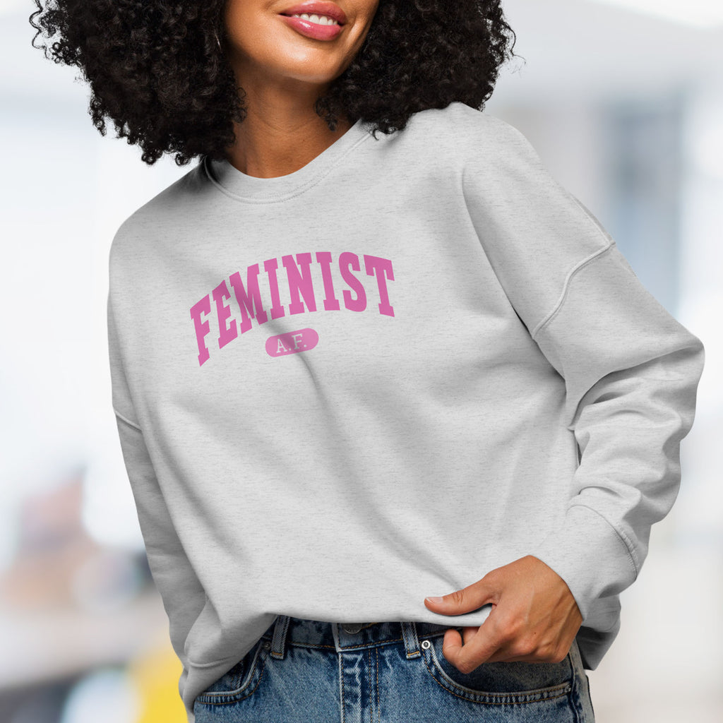 Front view of female model wearing grey sweatshirt with pink text that reads “Feminist A.F.” in a collegiate style.