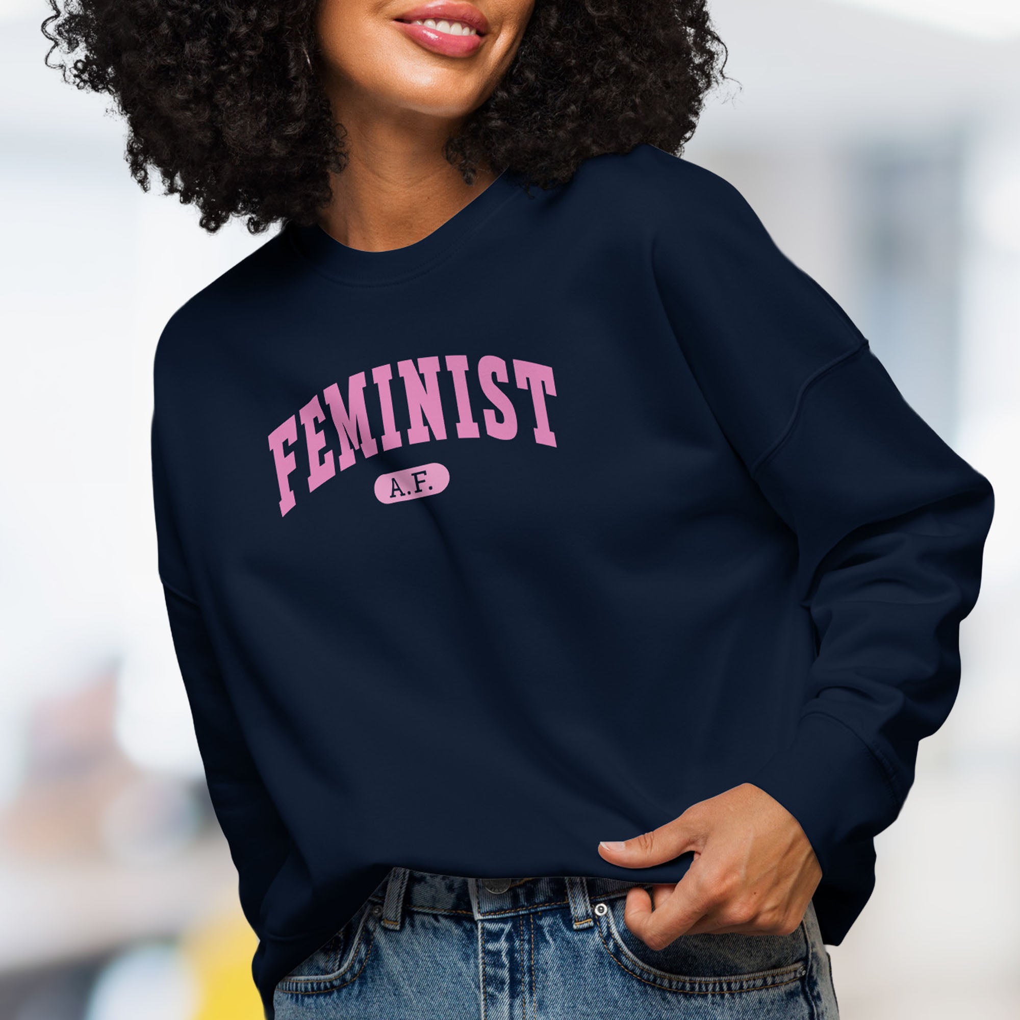 Front view of female model wearing navy sweatshirt with pink text that reads “Feminist A.F.” in a collegiate style.
