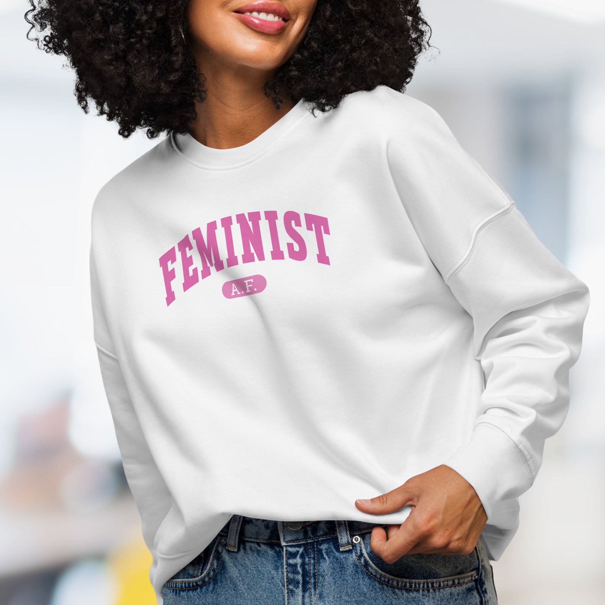 Front view of female model wearing white sweatshirt with pink text that reads “Feminist A.F.” in a collegiate style.