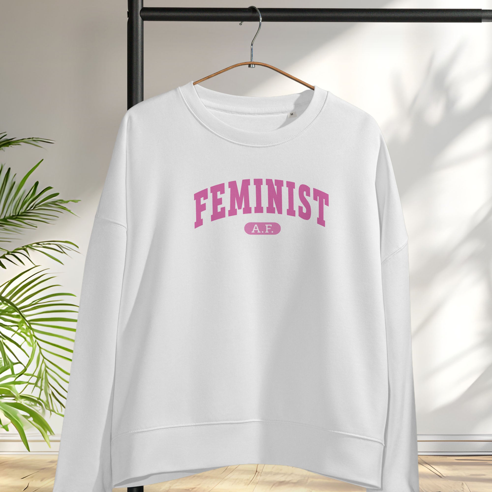 Hanging white sweatshirt with pink text that reads “Feminist A.F.” in a collegiate style.