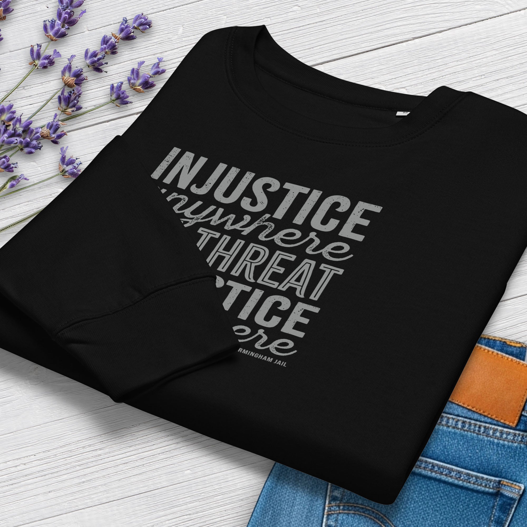 Folded black sweatshirt with bold text reading “Injustice anywhere is a threat to justice everywhere” quote from Martin Luther King Jr, MLK Jr, Letters from Birmingham jail — civil rights protest apparel for activists. Styled with jeans and flowers.